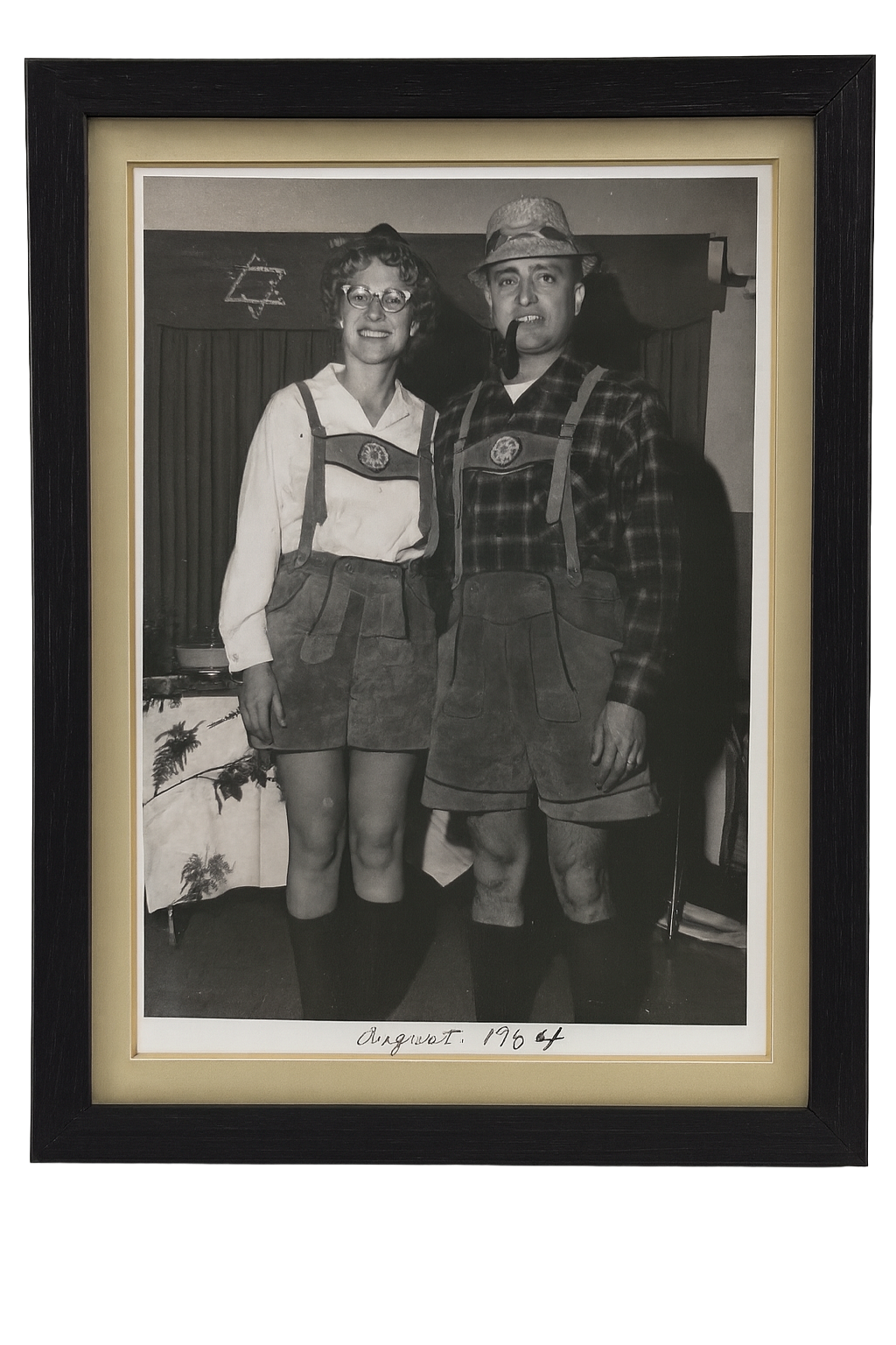 Black and white photograph of a woman and man dressed in lederhosen costumes, standing indoors with a curtain and Star of David in the background, dated August 1964.