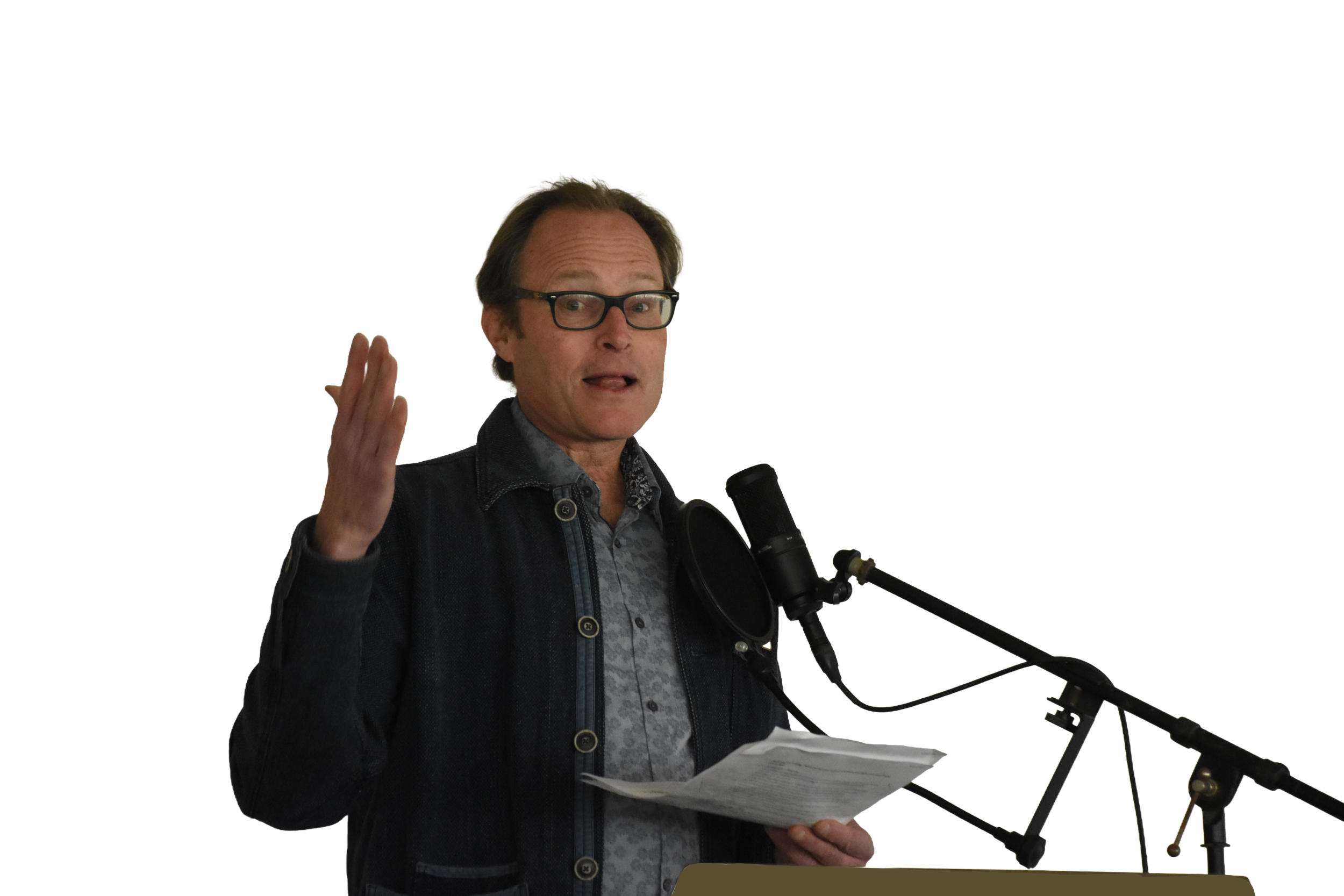 A man with glasses and dark hair speaking into a microphone while holding papers, gesturing with his right hand.