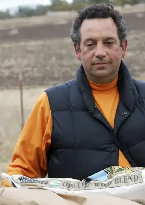 A man with curly dark hair wearing an orange shirt and a navy puffy vest outdoors, with a blurry dirt field in the background.