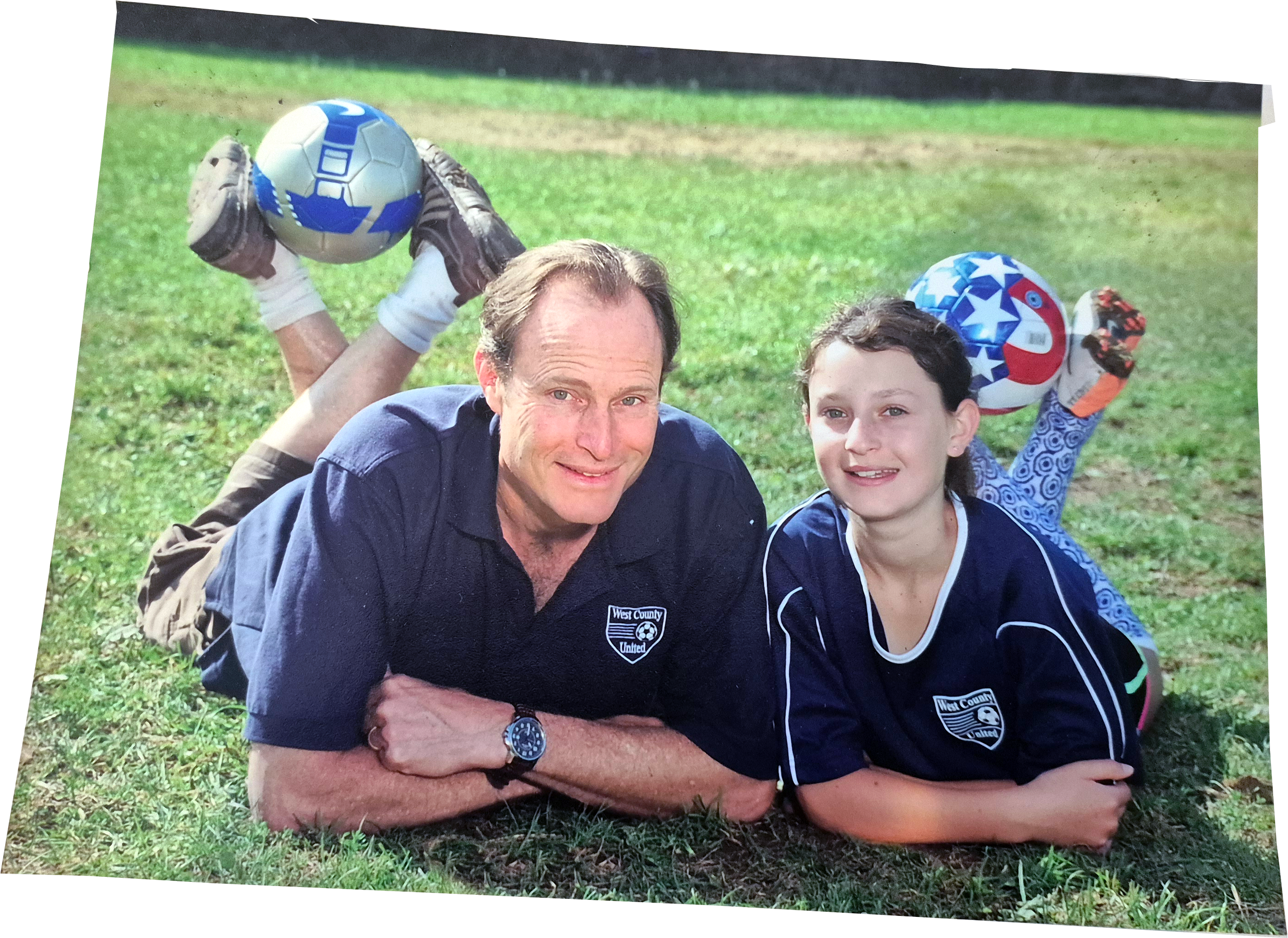 A man and a girl in soccer jerseys lying on grass, smiling, with two children in goalkeeper gear holding soccer balls behind them.