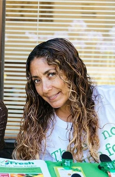A woman with long curly hair smiling at the camera during an outdoor event, sitting at a table with pamphlets and small bottles.