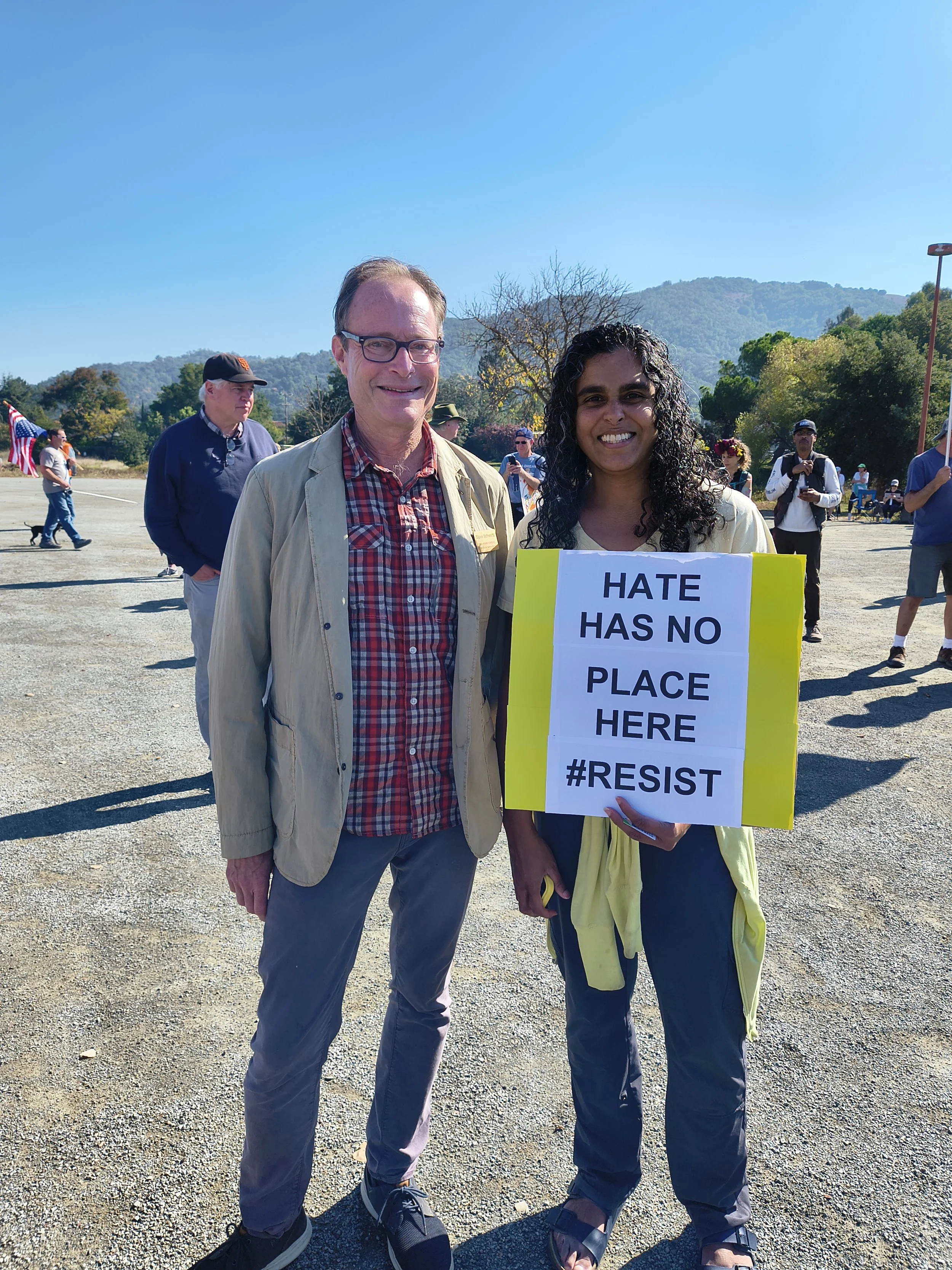 A woman holding a sign that reads 'HATE HAS NO PLACE HERE #RESIST' standing next to a man on a sunny day at an outdoor event, with several people in the background.