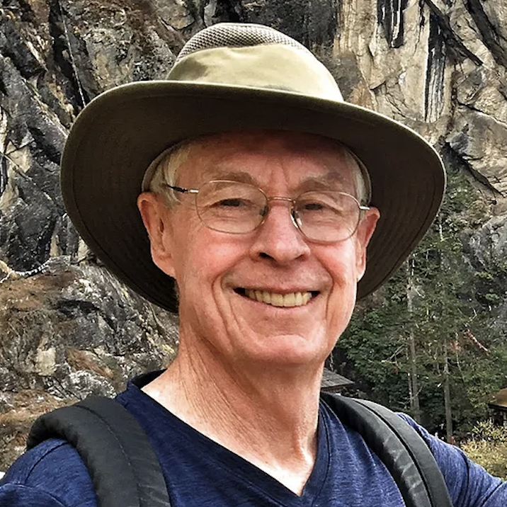 A smiling older man wearing glasses, a wide-brimmed hat, a navy blue shirt, and a backpack, standing outdoors in front of rocky terrain with trees.