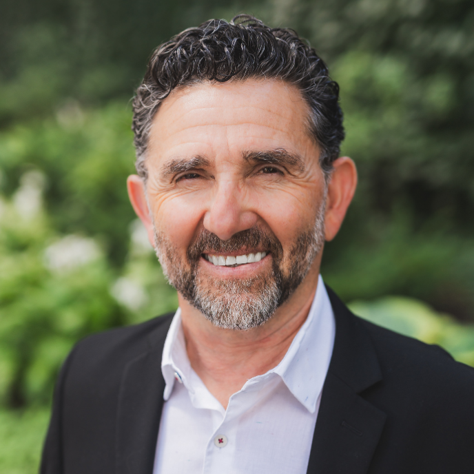 A smiling middle-aged man with dark, curly hair and a beard, wearing a black blazer and white shirt, standing outdoors with a blurred green background.