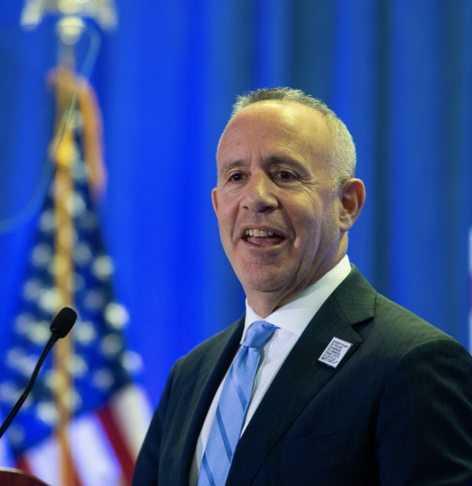 A man in a suit and light blue tie speaking at a podium with an American flag in the background.