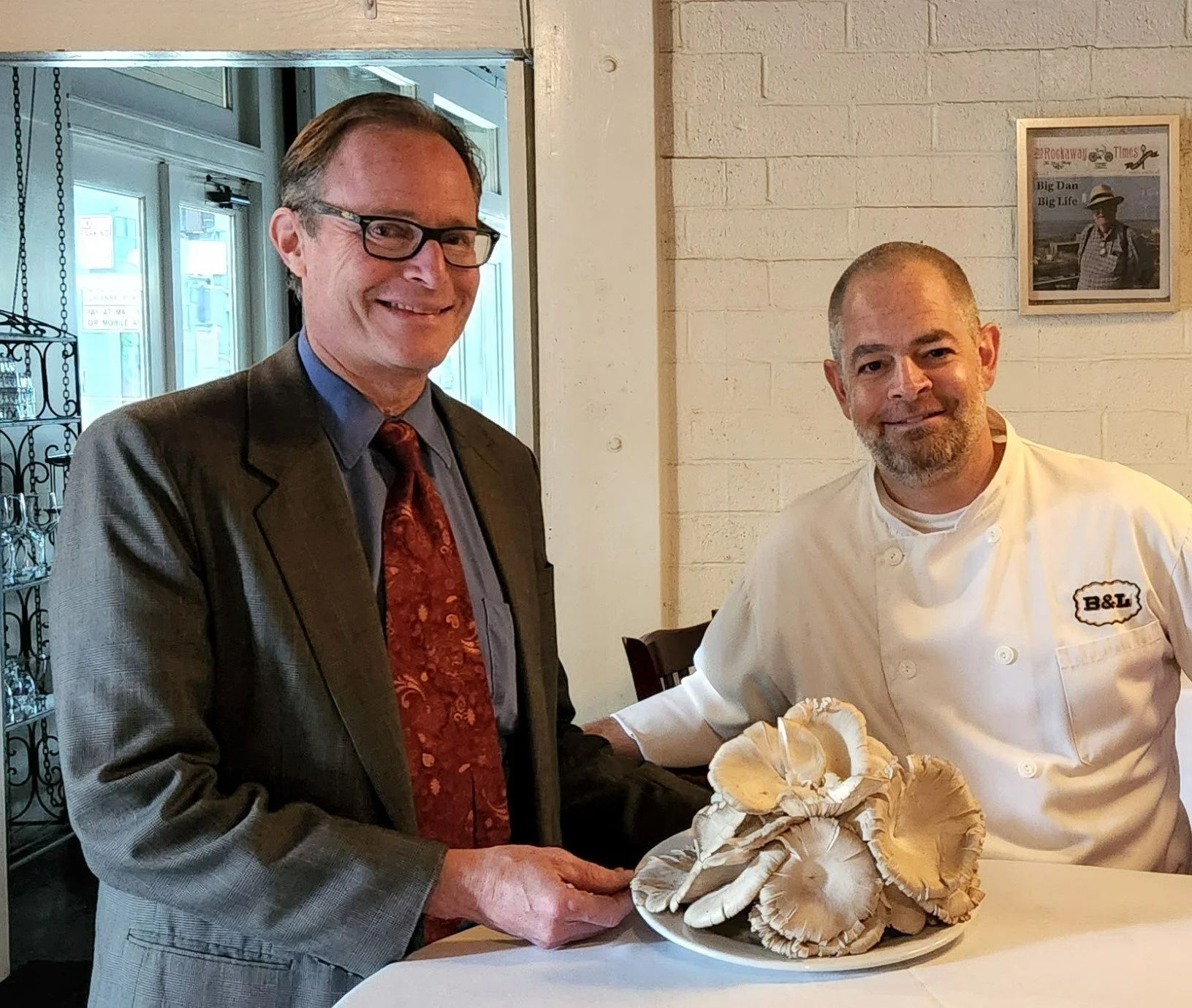 A man in a suit and a chef are standing next to each other at a table with a bowl of mushrooms. The man is holding a mushroom and both are smiling.