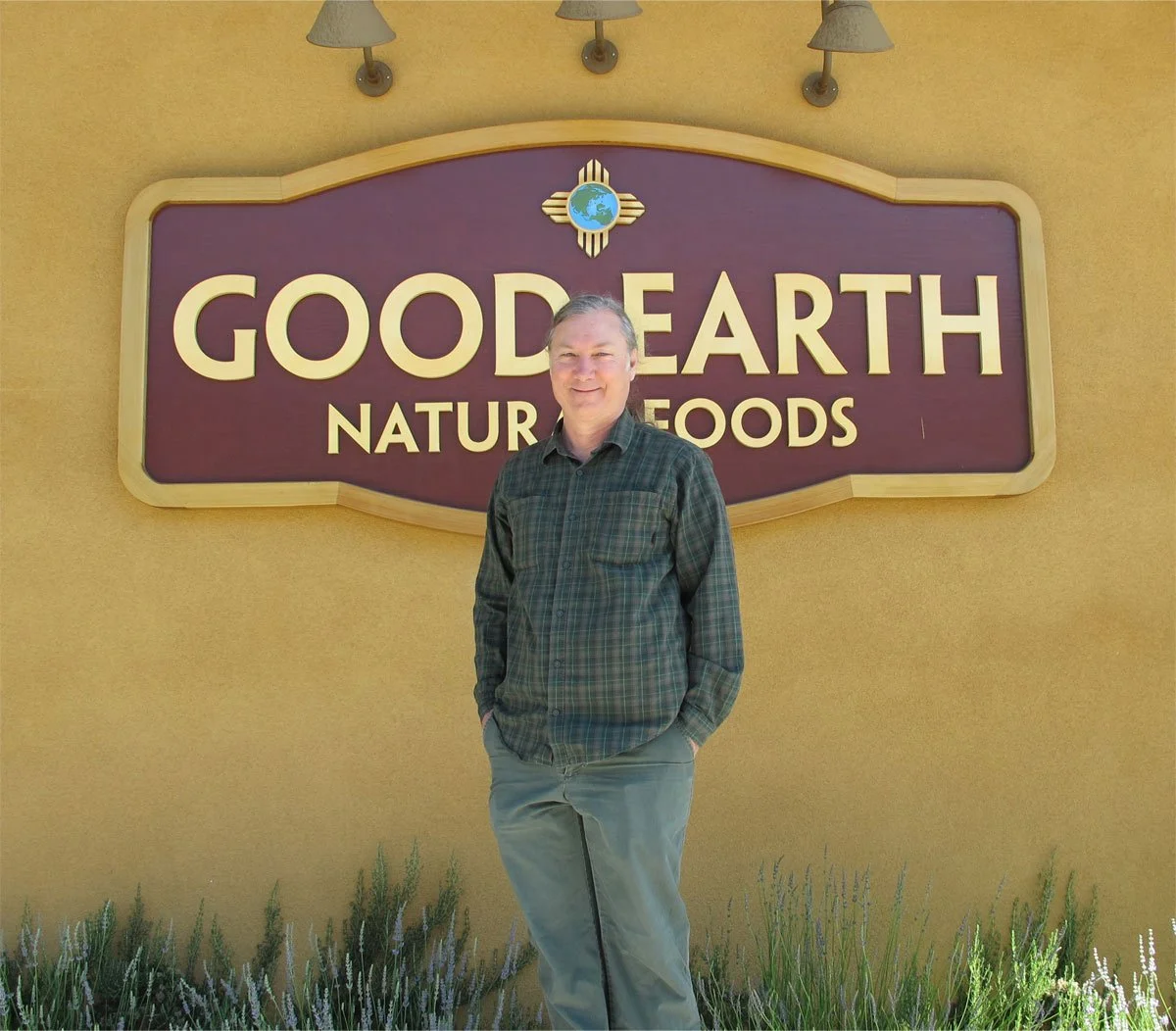 A man standing in front of a sign that reads "Good Earth Natural Foods" on a yellow wall with three light fixtures above the sign and plants at the base.
