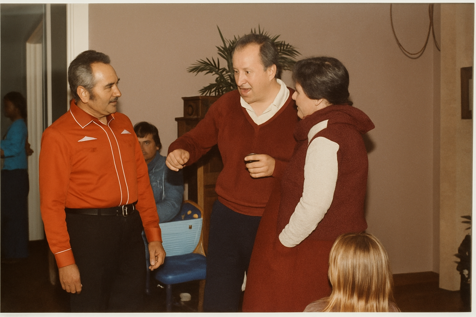 Three people are having a conversation in a room, one man in a red shirt and two others, a man and a woman, in casual clothing. There is a young girl with red hair sitting nearby, and other people are visible in the background.