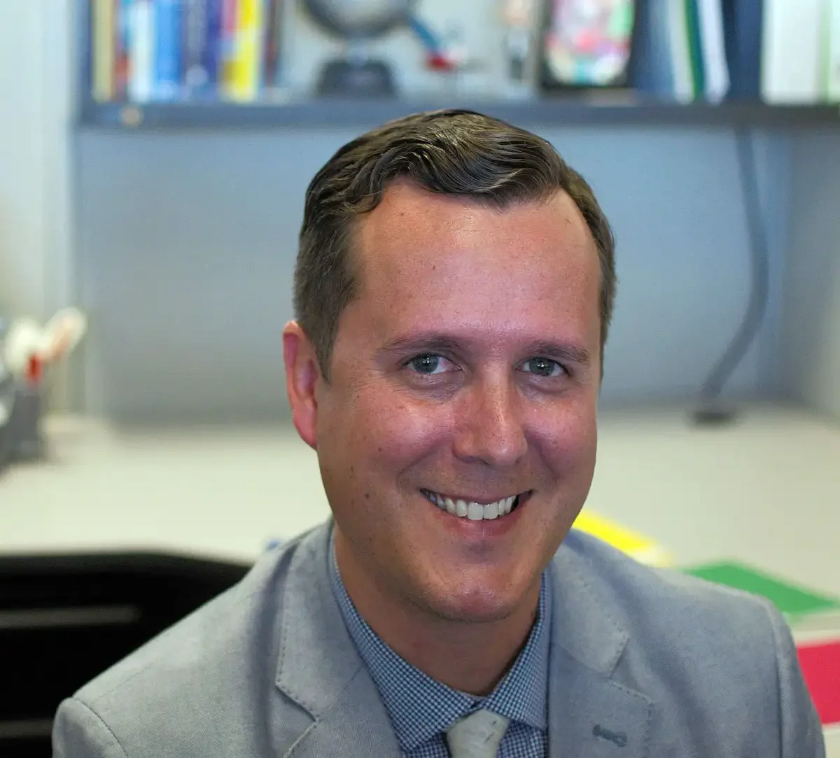 A man in a gray suit smiling in a professional office setting with shelves of books and folders in the background.