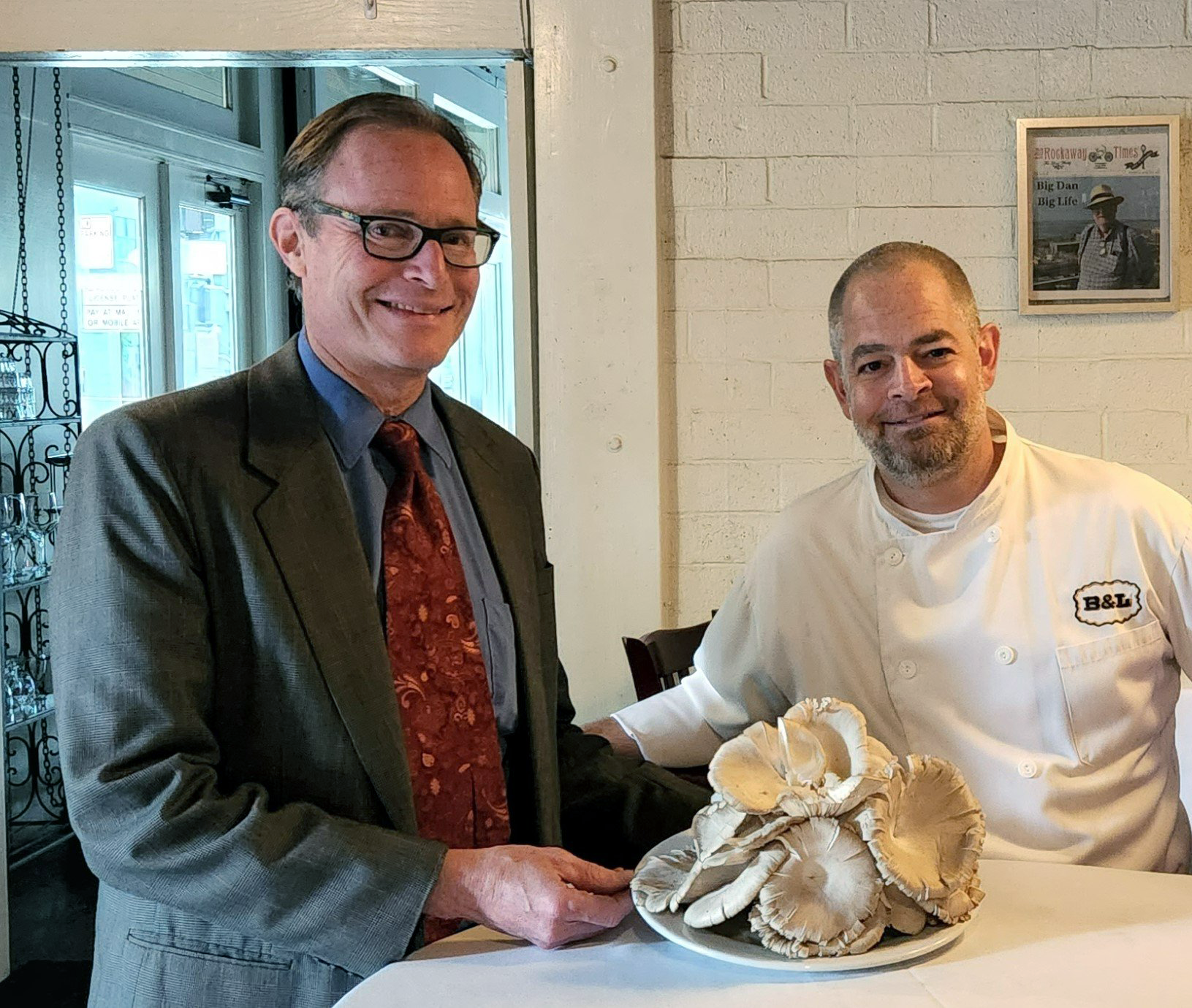 Two men, one in a suit and glasses, the other in a chef's uniform, standing at a table with a plate of large mushrooms.