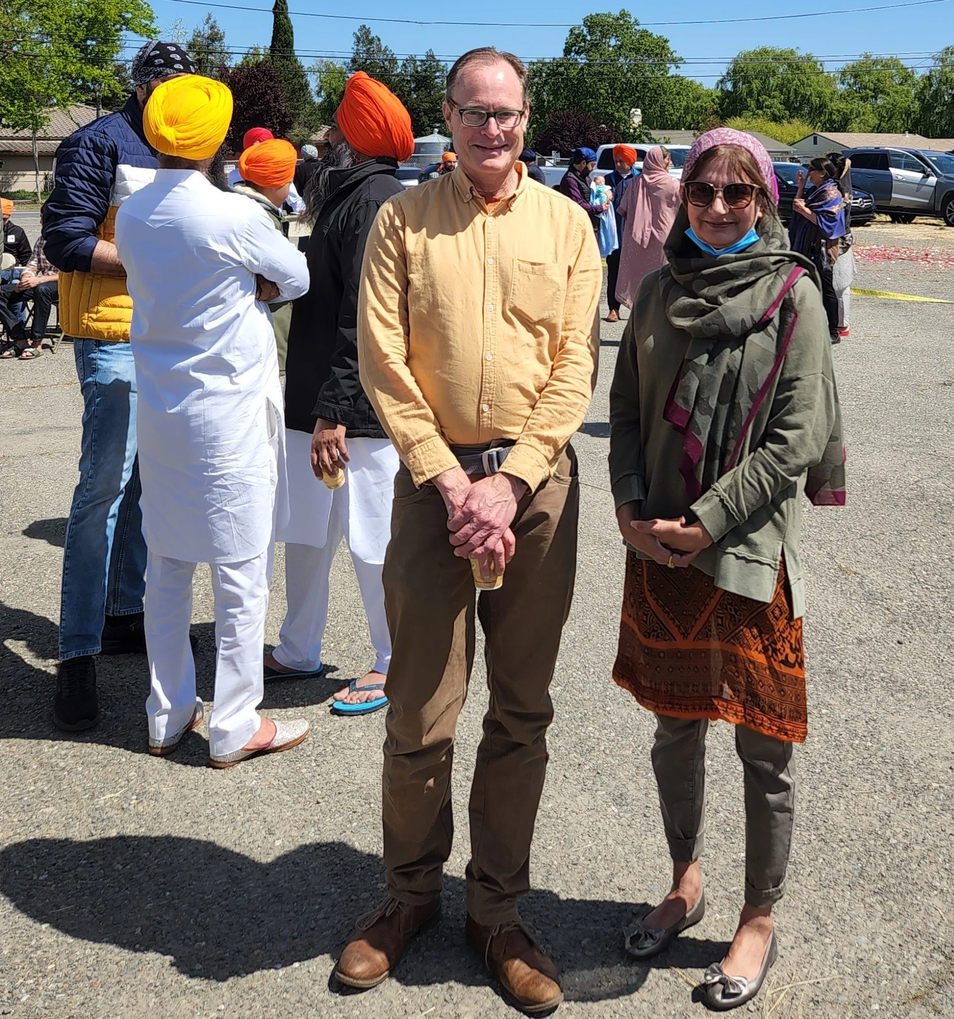 Two people standing outdoors at a gathering, surrounded by others, with trees, parked cars, and a blue sky in the background.