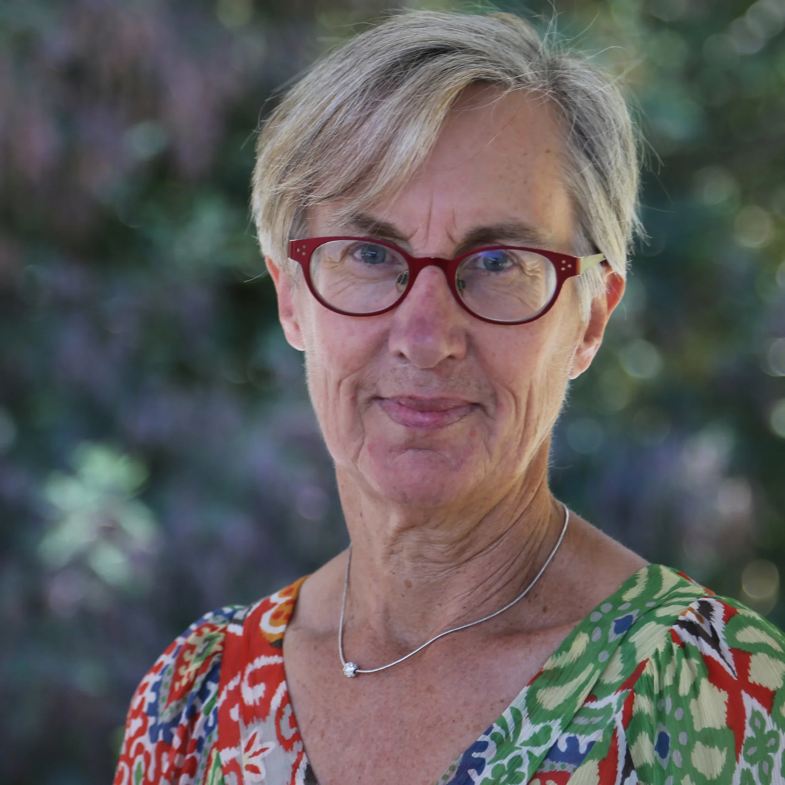 A portrait of an older woman with short gray hair, red glasses, wearing a colorful patterned blouse and a delicate silver necklace, standing outdoors with a blurred background of trees and greenery.