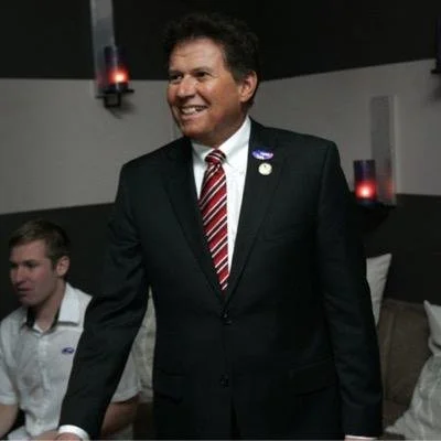 Man in a suit smiling at a social gathering with others seated around.