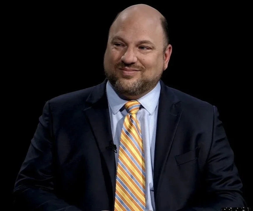A man in a dark suit with a light blue shirt and a yellow striped tie, smiling and looking to the right, against a black background.