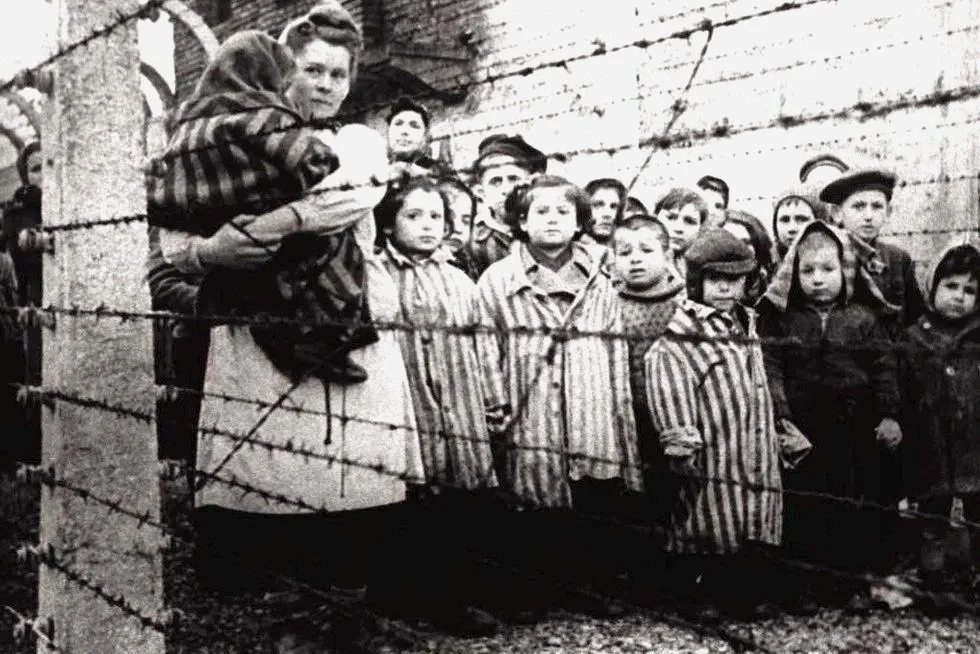 Group of children standing behind a barbed wire fence, some with adults, in a historical black and white photograph.