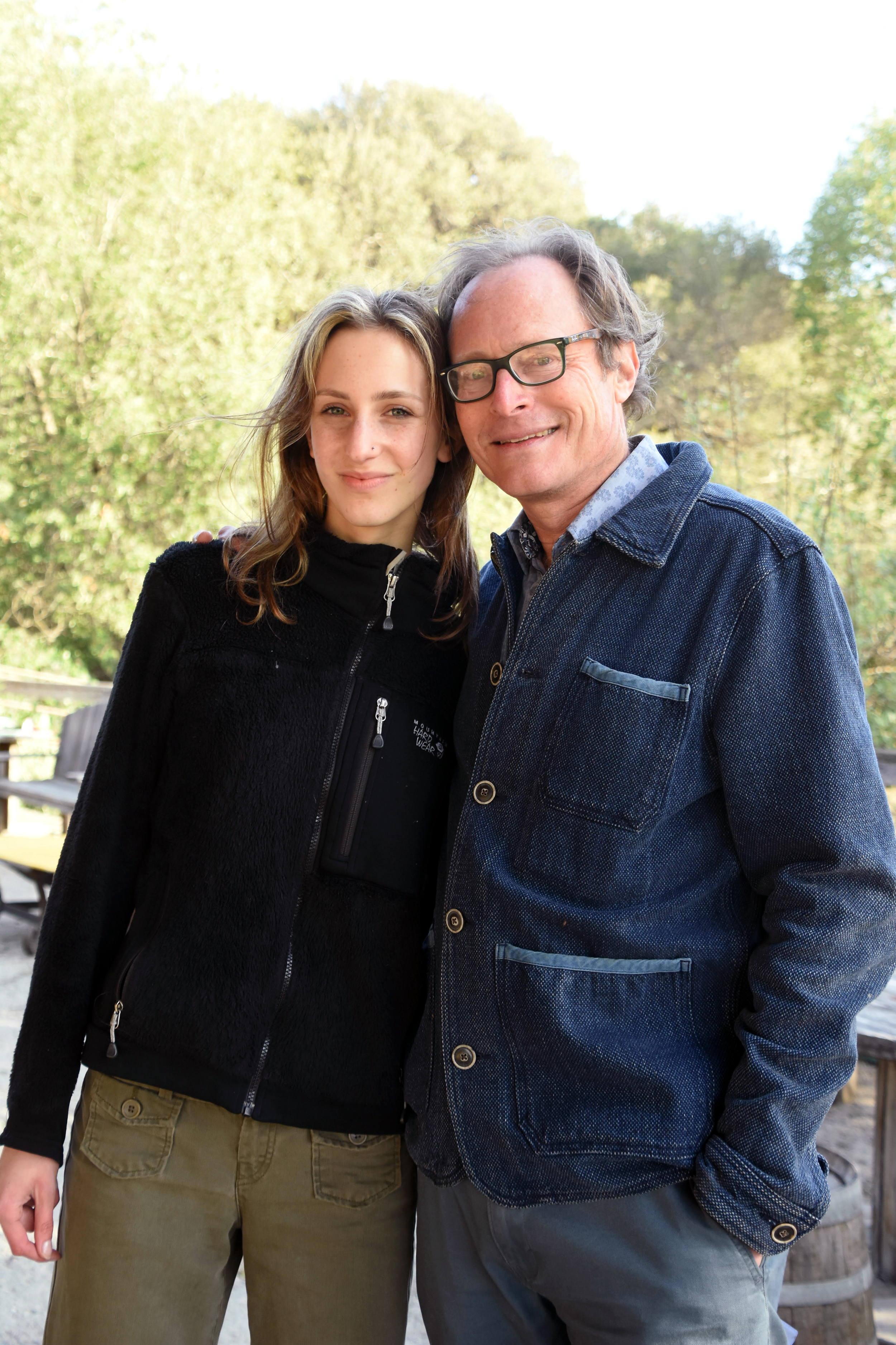 A young woman and an older man standing together outdoors, smiling, with trees and a clear sky in the background.