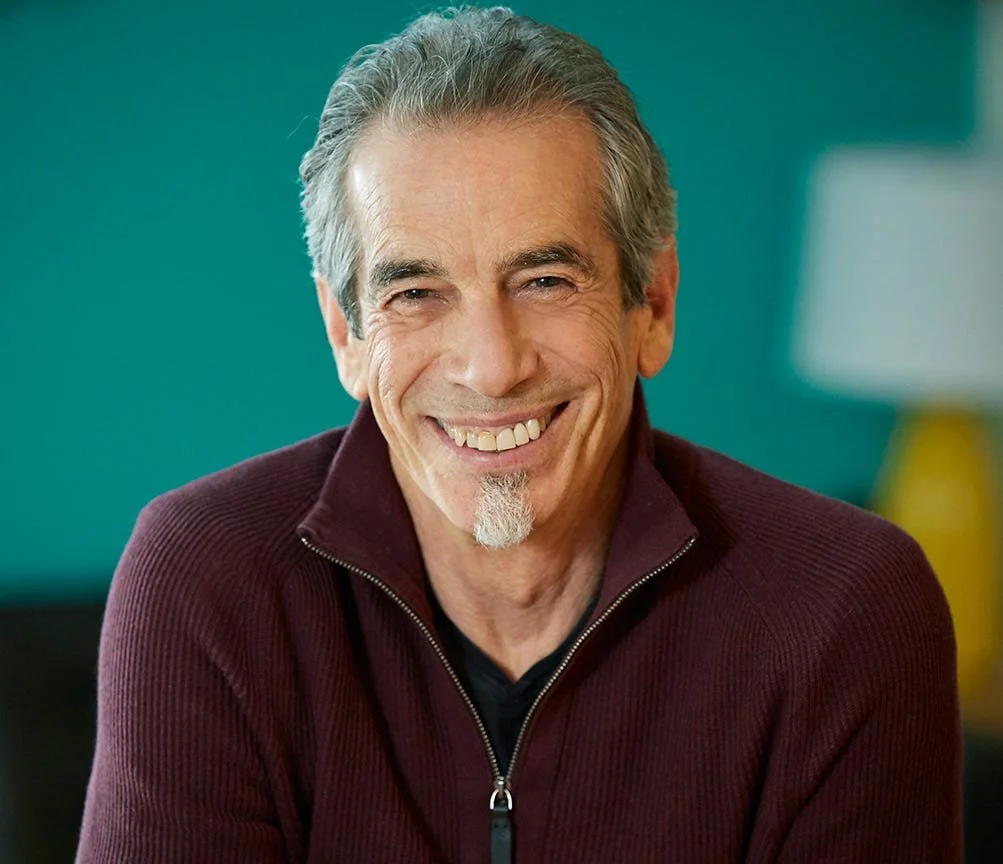A smiling middle-aged man with gray hair and a goatee, wearing a maroon zip-up sweater, in a well-lit room with a blurred teal background.