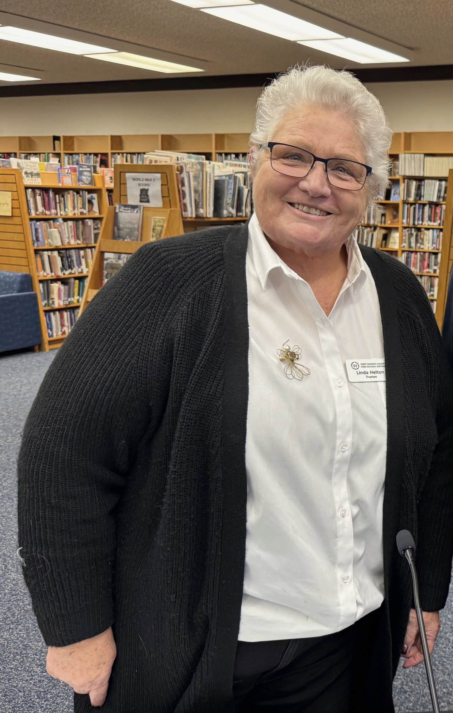 An older woman with short white hair, glasses, and a warm smile, standing in a library. She is wearing a white button-up shirt with a pin and a black cardigan. The background shows bookshelves filled with books.
