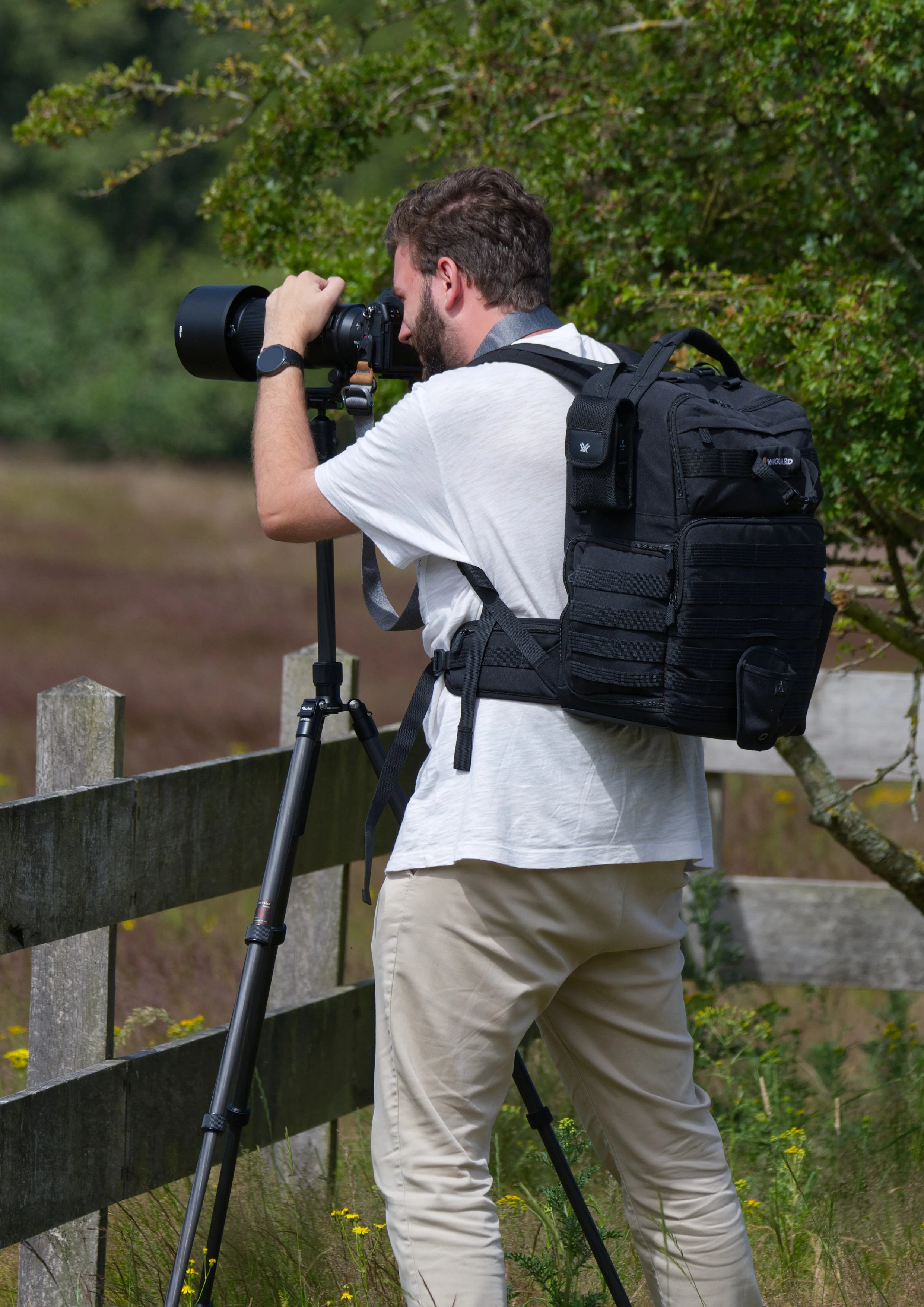 Man met een camera op een statief, buiten, voor een houten hek, omringd door groen**