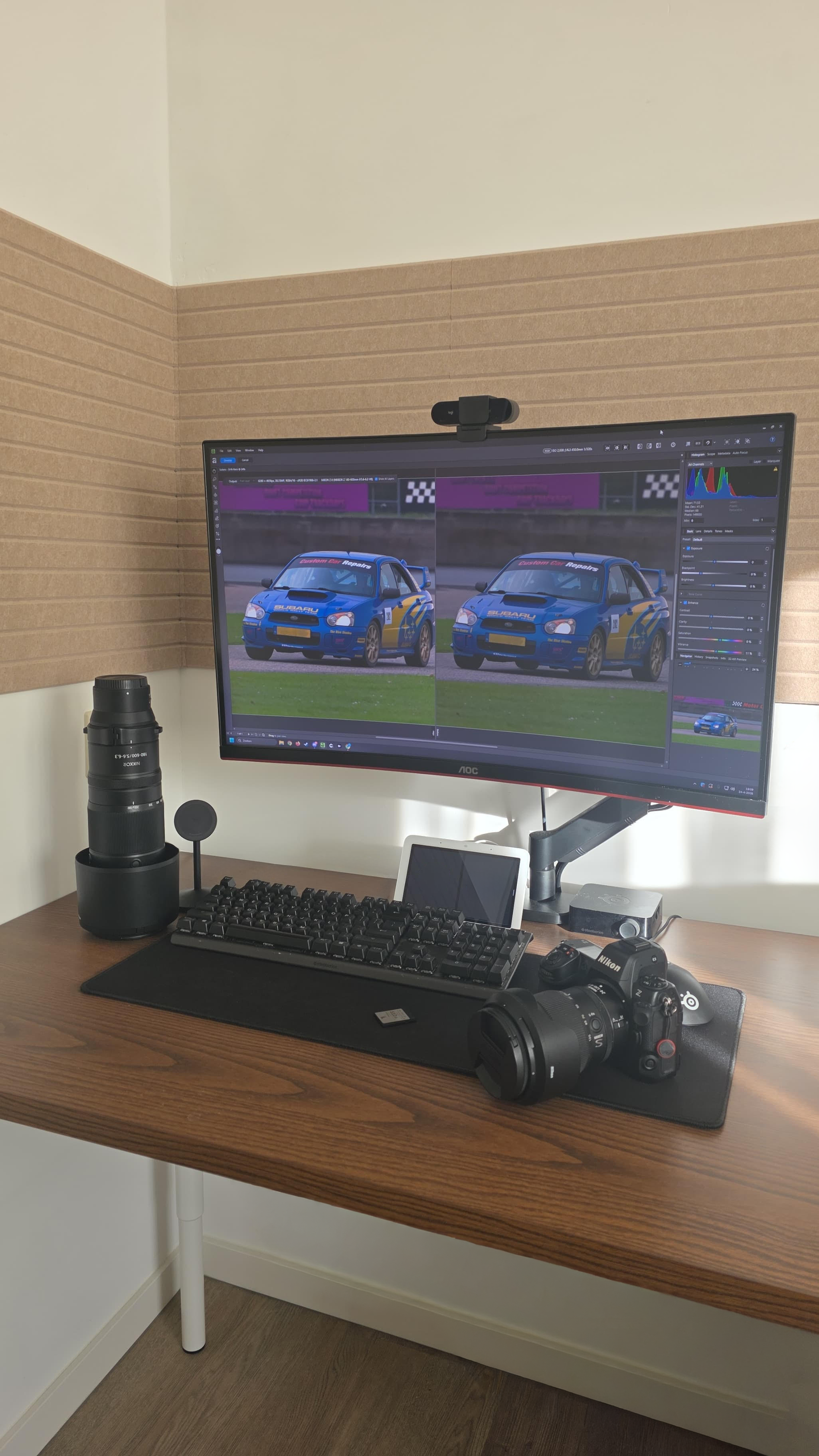Een werkplek met een monitor die een fotobewerkingsprogramma toont met twee foto's van een blauwe rallyauto, een grote telelens, een toetsenbord, een muis, een smartphone, een Nikon camera en andere digitale apparatuur. De werkplek bevindt zich tegen een muur met een deels versoberde groene en beige kleur.