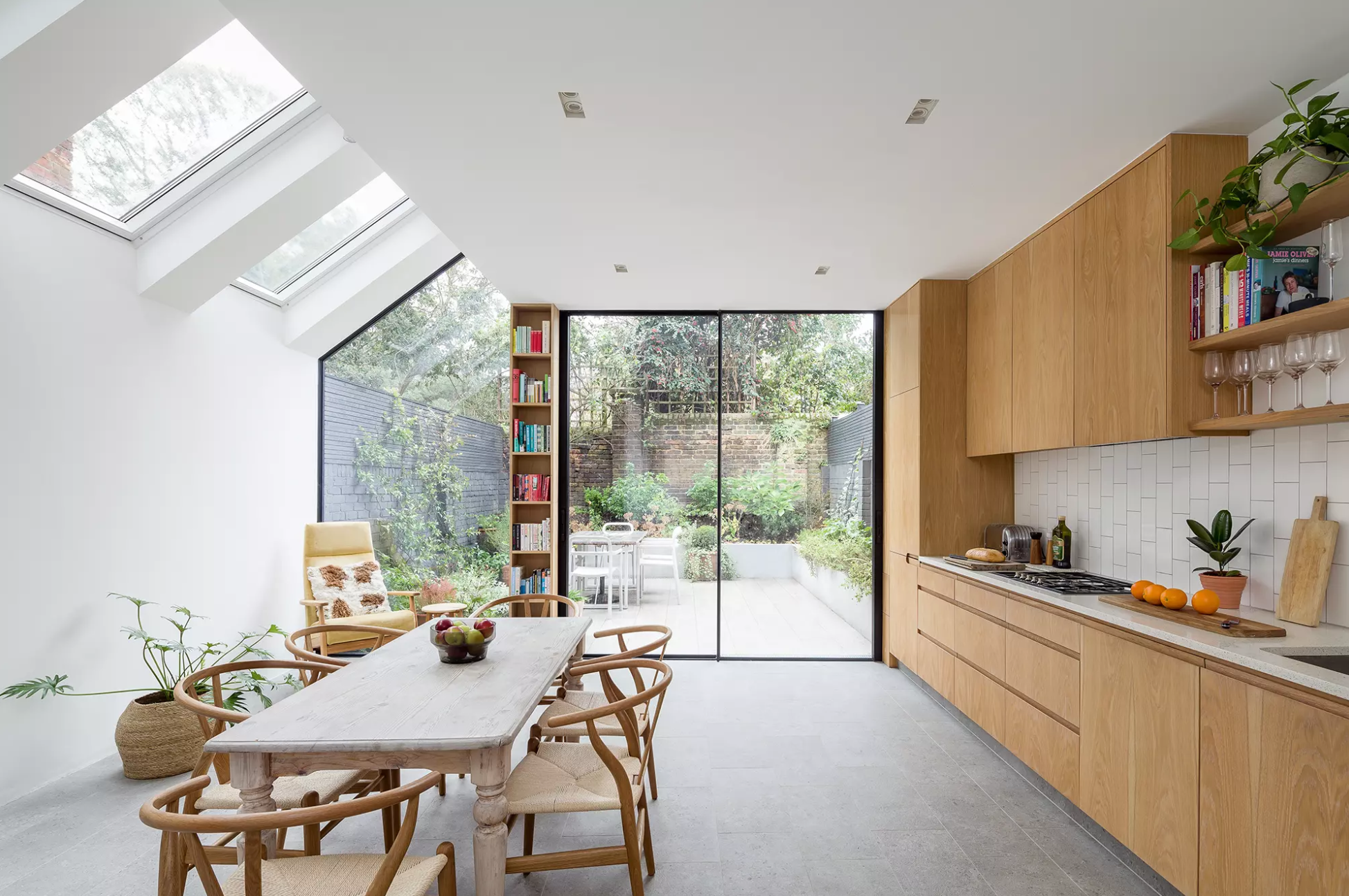 Modern London kitchen extension by design and build architects, with large skylights, floor-to-ceiling glass doors, bespoke wooden cabinetry, and a lush garden view.
