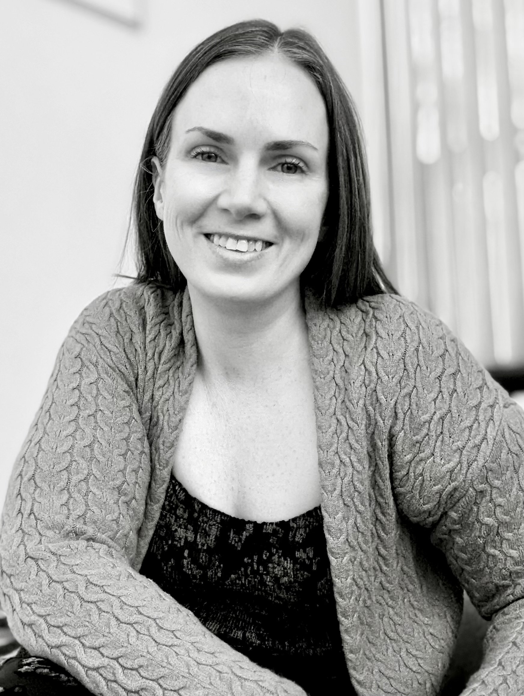 Black and white photo of a smiling trauma therapist with straight dark hair, wearing a patterned top and a textured cardigan, sitting indoors with a blurred background.