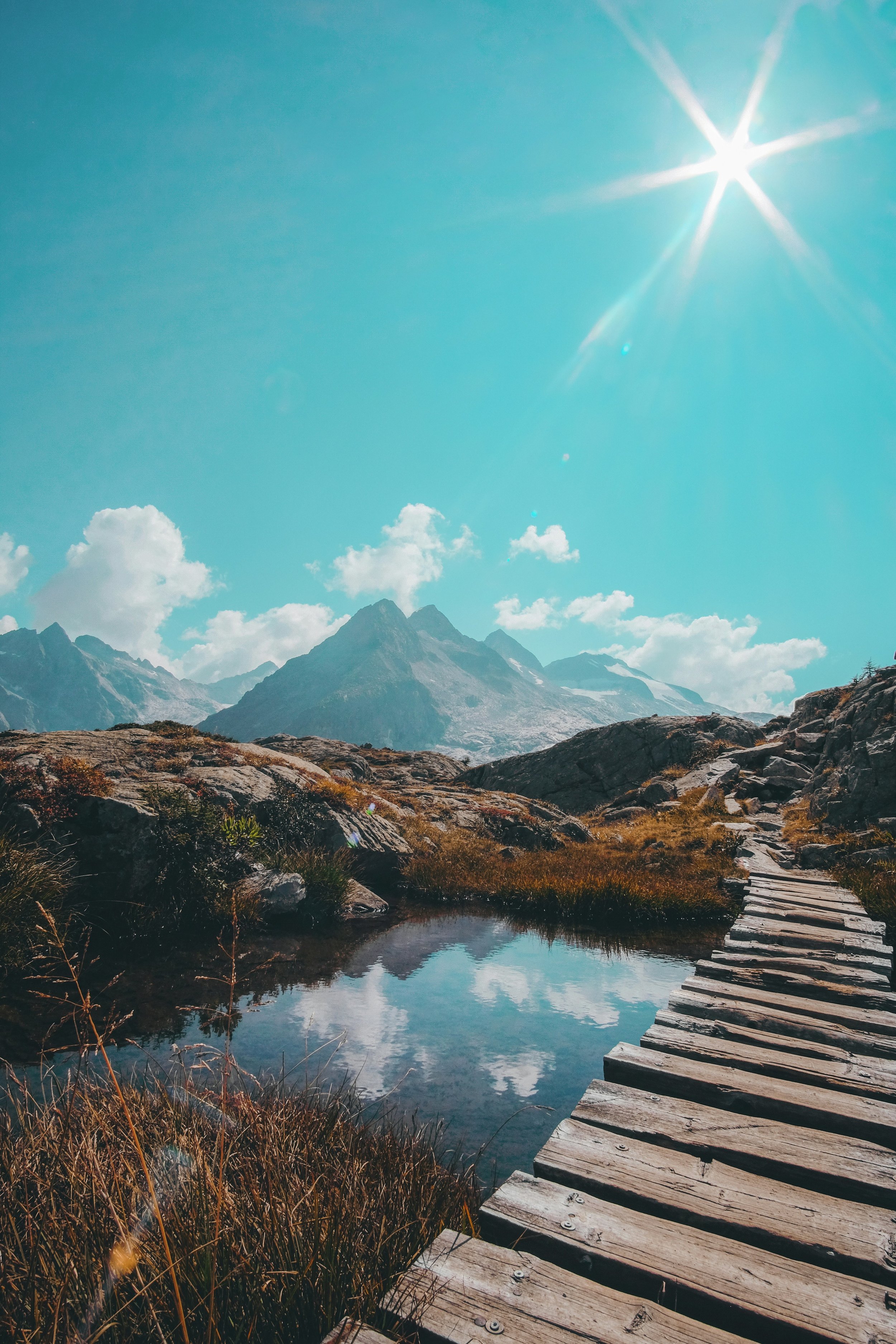 Mountain landscape with trail leading to mountains, small pond reflecting clouds, bright sun in blue sky with some clouds.