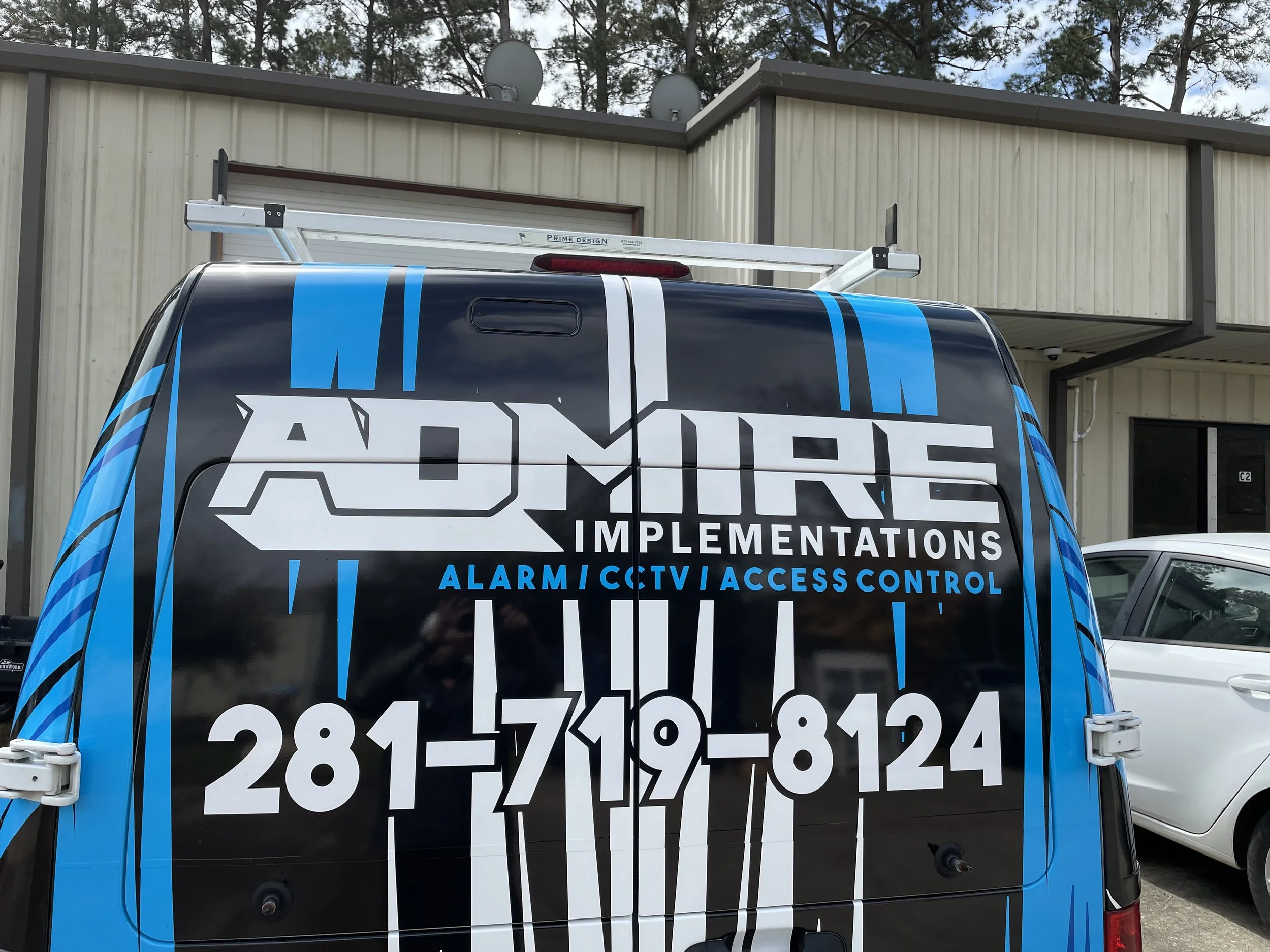 Close-up of a black van with a large logo and contact information for ADMSI, a security system installation company, with a building and other vehicles in the background.
