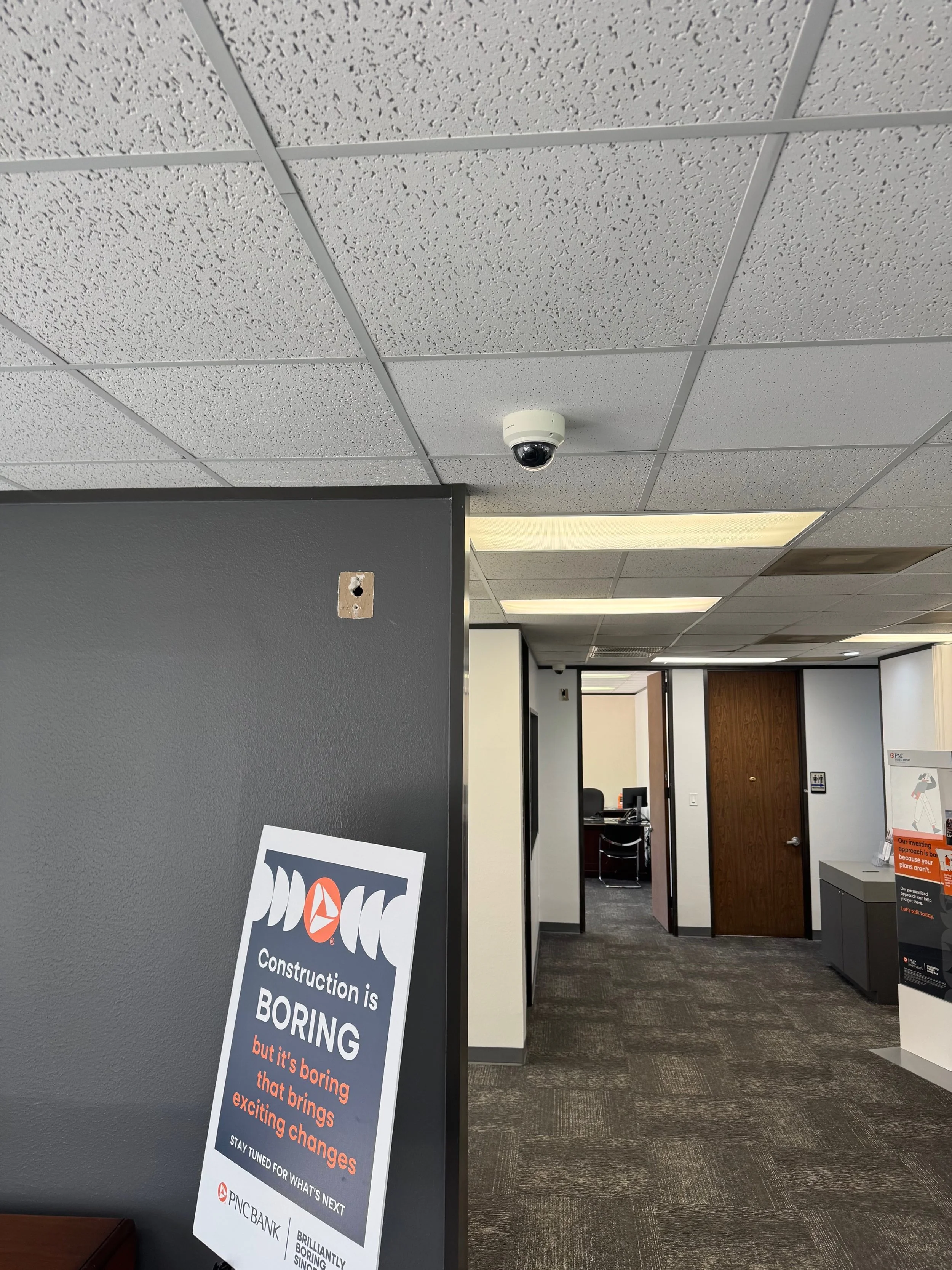 An interior shot of an office with a gray wall, a security camera on the ceiling, and a sign that reads 'Construction is boring but it’s boring that brings exciting changes.' There are wooden doors, office furniture, and a ceiling with fluorescent li