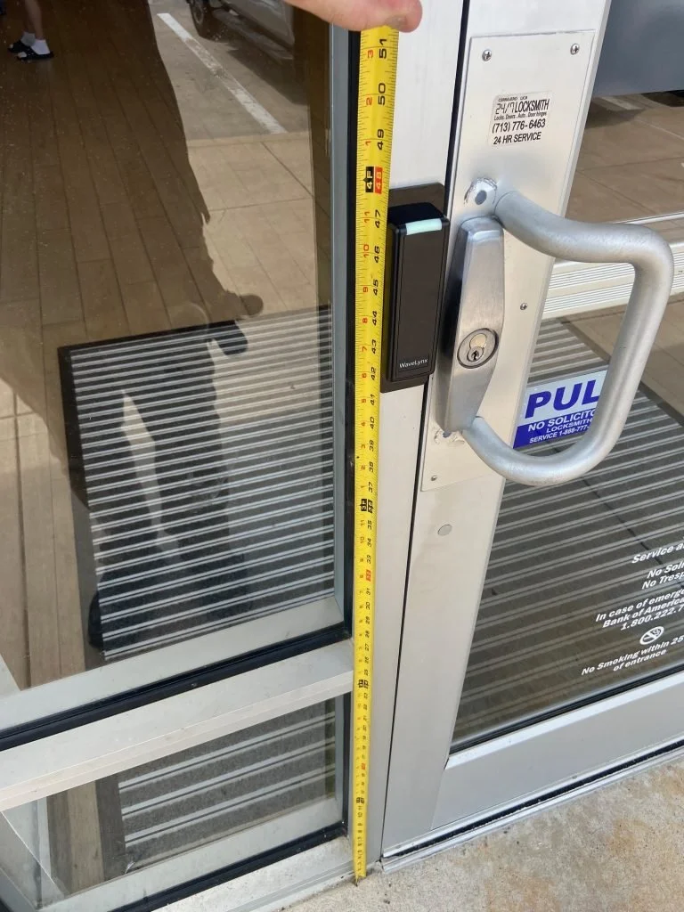 Close-up of a glass entrance door with a measuring tape indicating the door frame is approximately 50 inches tall. The door has a handle, a lock, and a security card scanner.