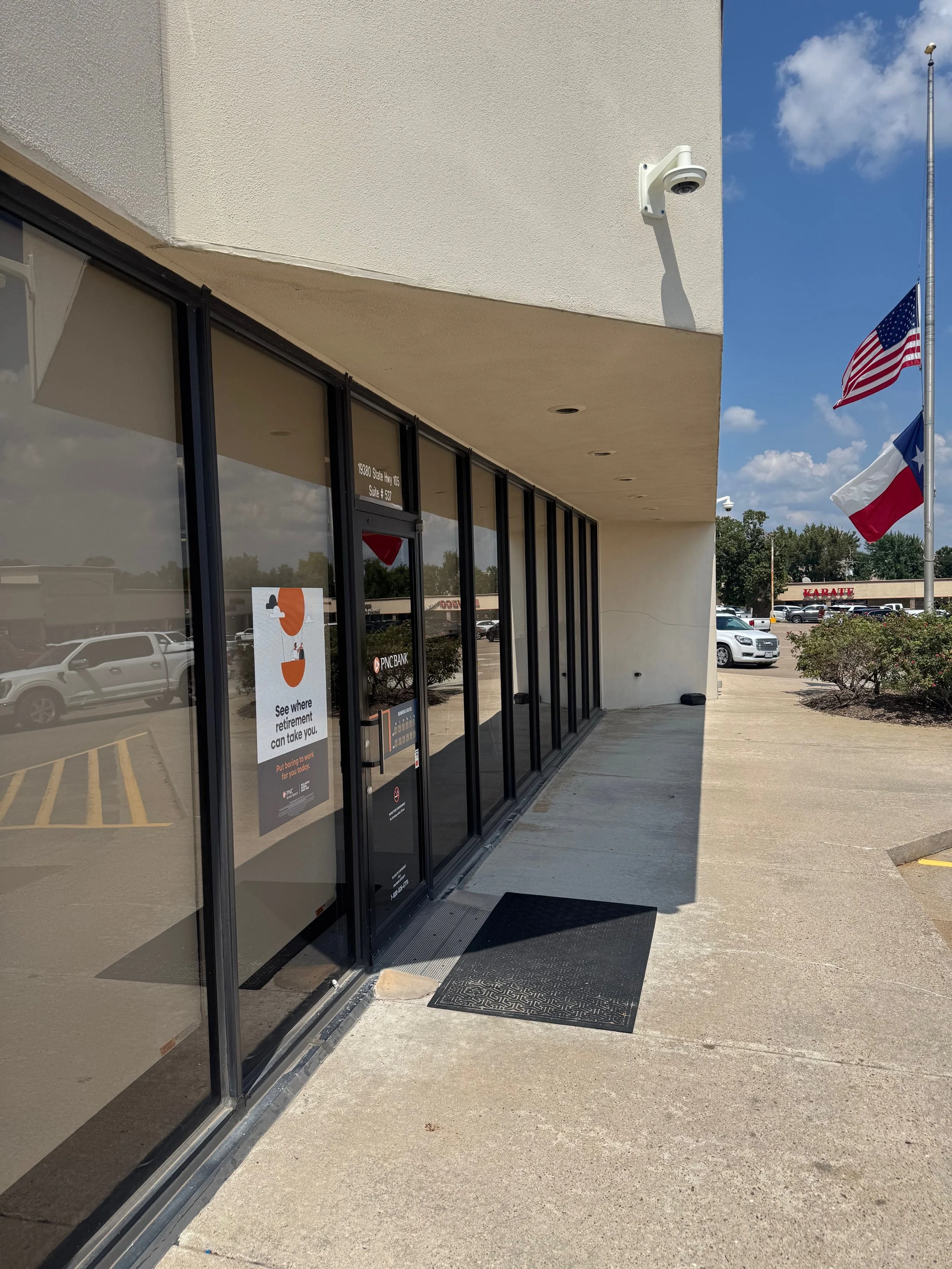 Exterior of a bank branch with a glass door and window, security cameras, and flags of the United States and Texas, with a parking lot and cars in the background on a sunny day.