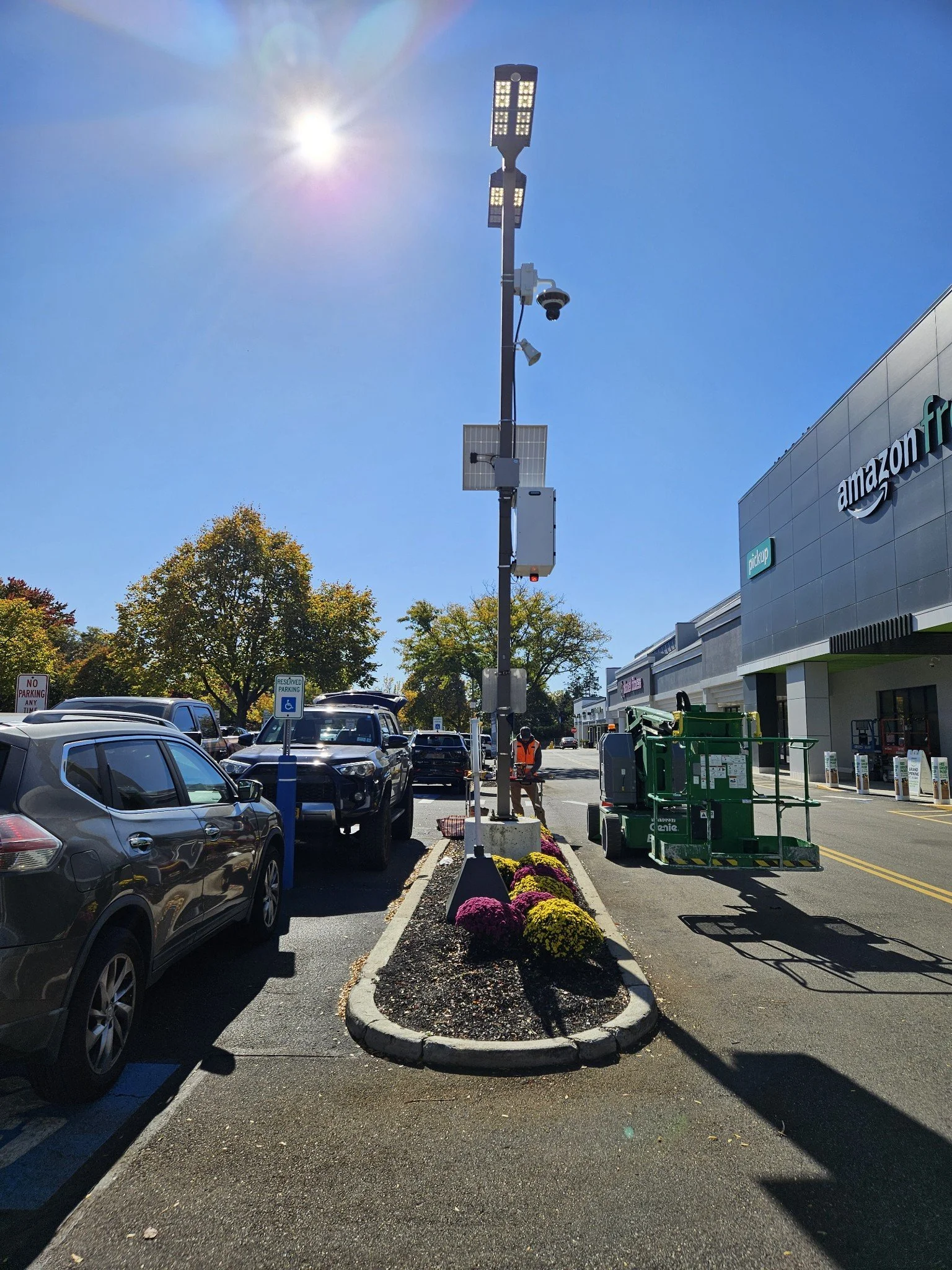 A parking lot outside an Amazon Fresh store with a landscaped median containing purple and yellow flowers, parking meters, and various vehicles, under a bright sunny sky.