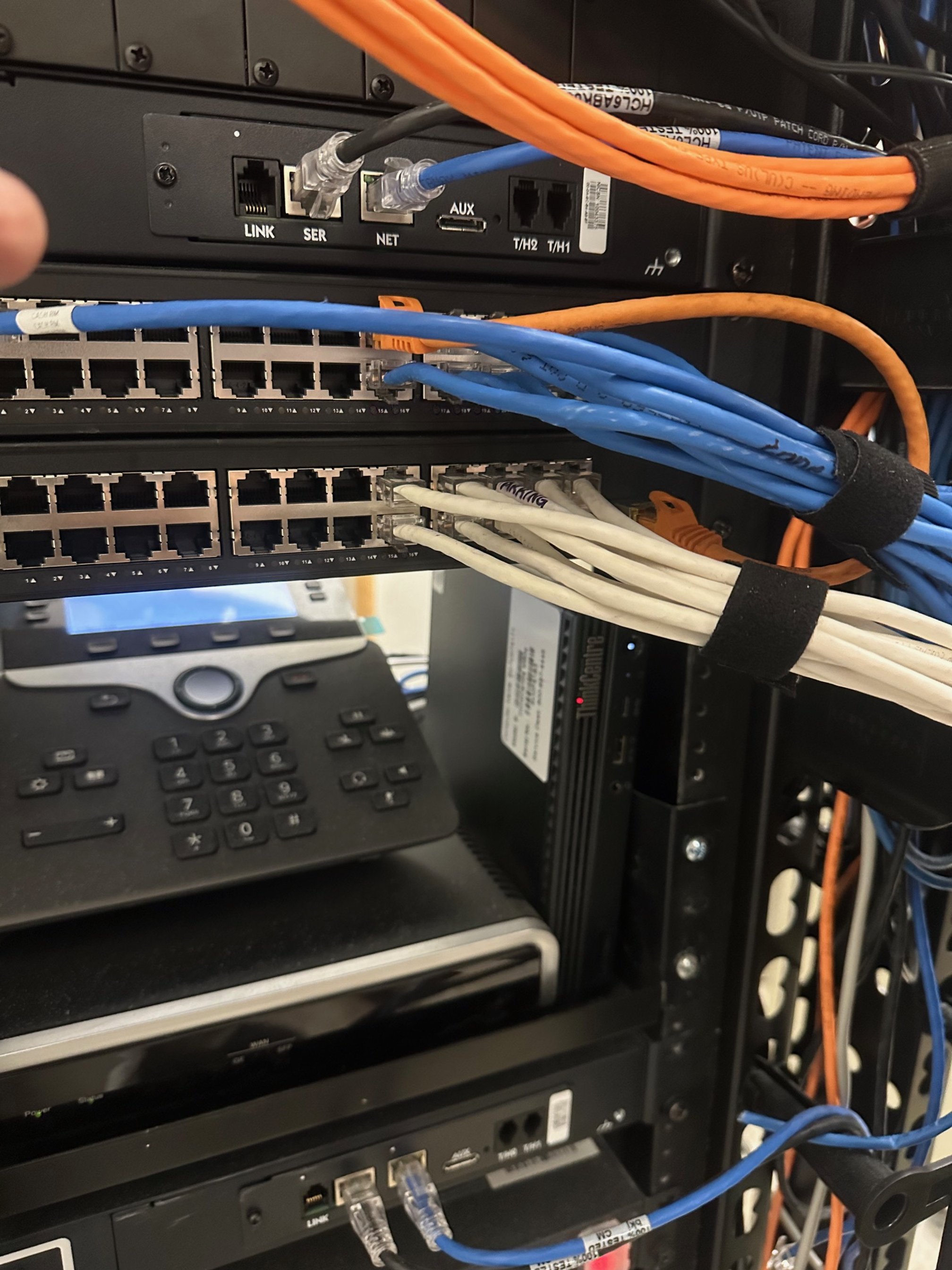 Close-up of a network server rack with multiple Ethernet cables connected to switches, and a black data device beneath a phone on the rack.