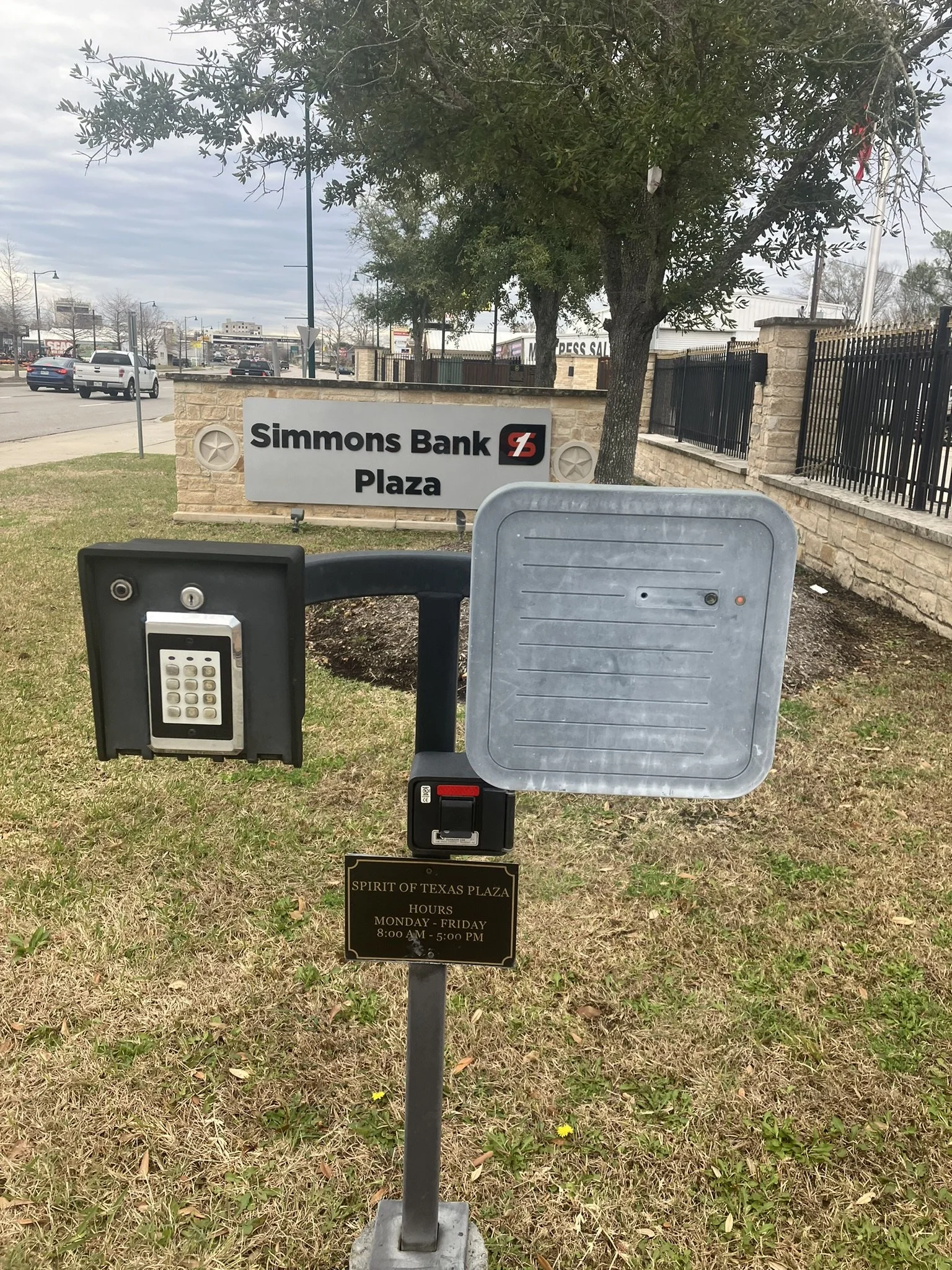 Sign for Simmons Bank Plaza behind a keypad access control panel and an empty curbside lockbox or drop box at Spirit of Texas Plaza, with a black plaque indicating hours of operation from Monday to Friday, 8:00 AM to 5:00 PM, on a grassy area with a 