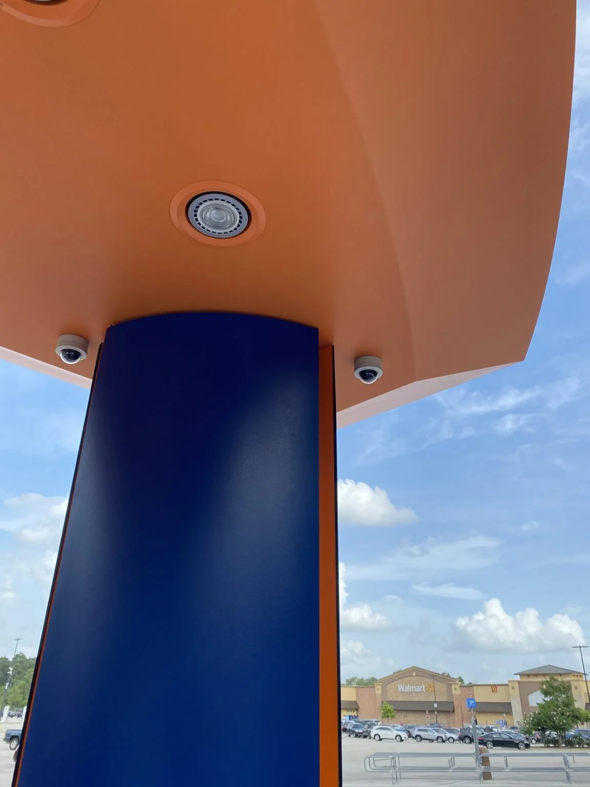 View of the ceiling with security cameras and a spotlight, part of the Walmart store exterior visible in the background under a partly cloudy sky.
