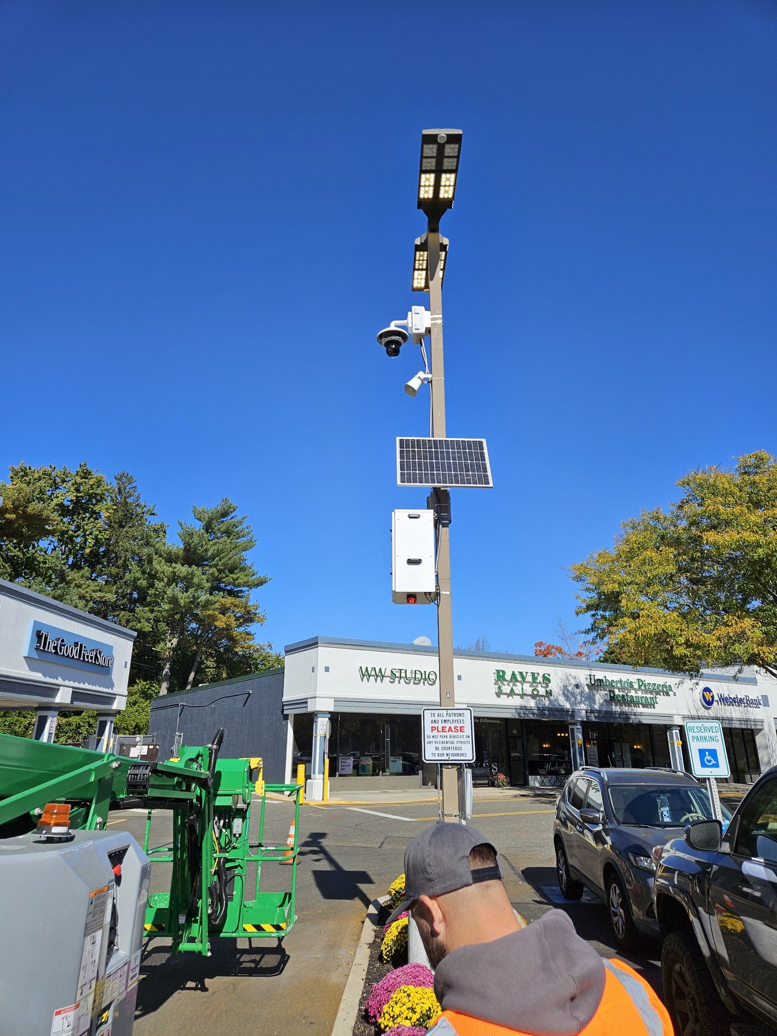 Street view featuring a tall pole with multiple surveillance cameras, a solar panel, and LED lights against a clear blue sky. In the foreground, there is a person with a baseball cap and an orange vest, along with parked cars and a strip mall with va
