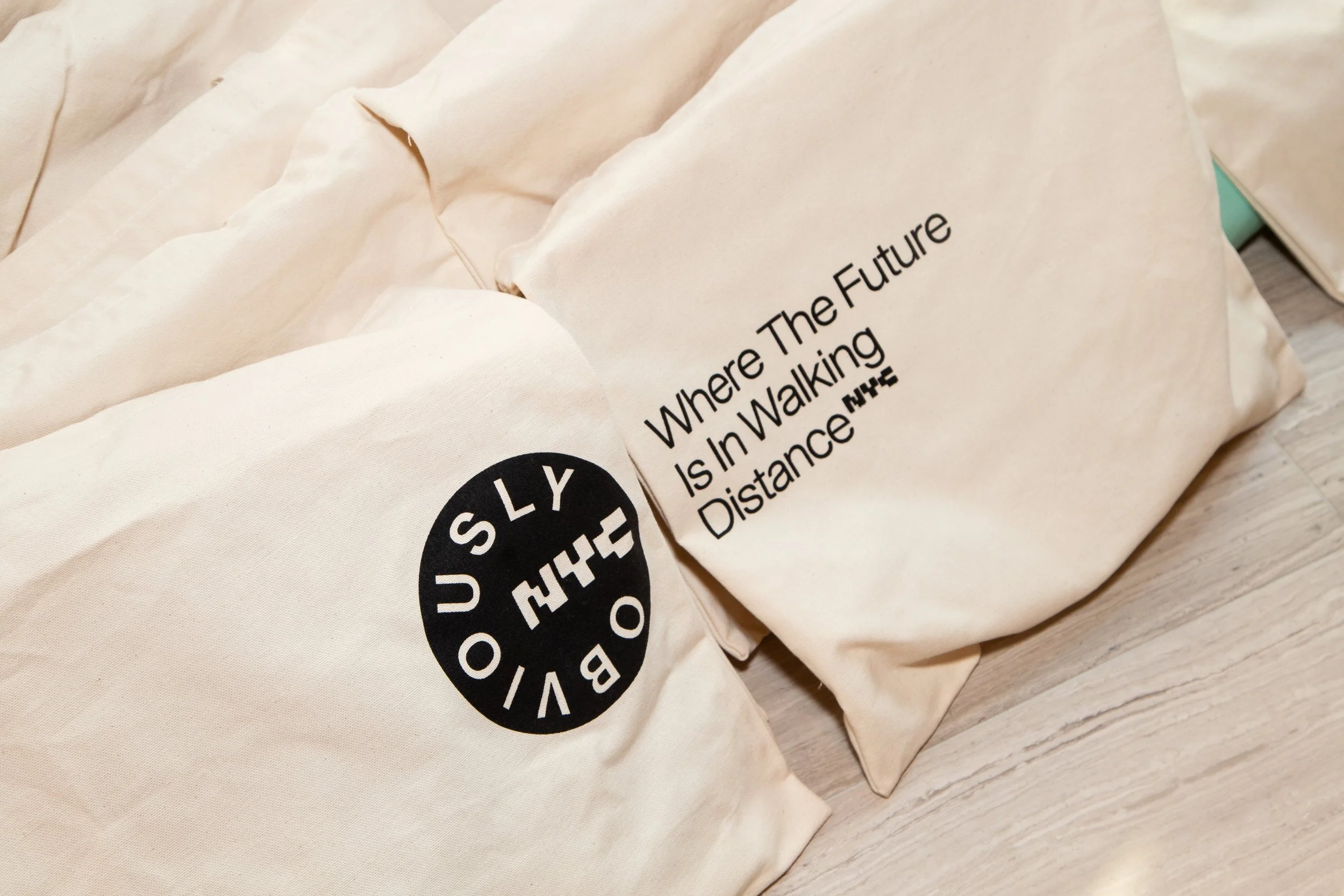 Two beige tote bags with black text, one reads 'OBVIOUSLY THE FUTURE IS IN WALKING DISTANCE' and the other has a black circular logo with 'NH THE' and 'OBVIOUSLY' written around the circle.
