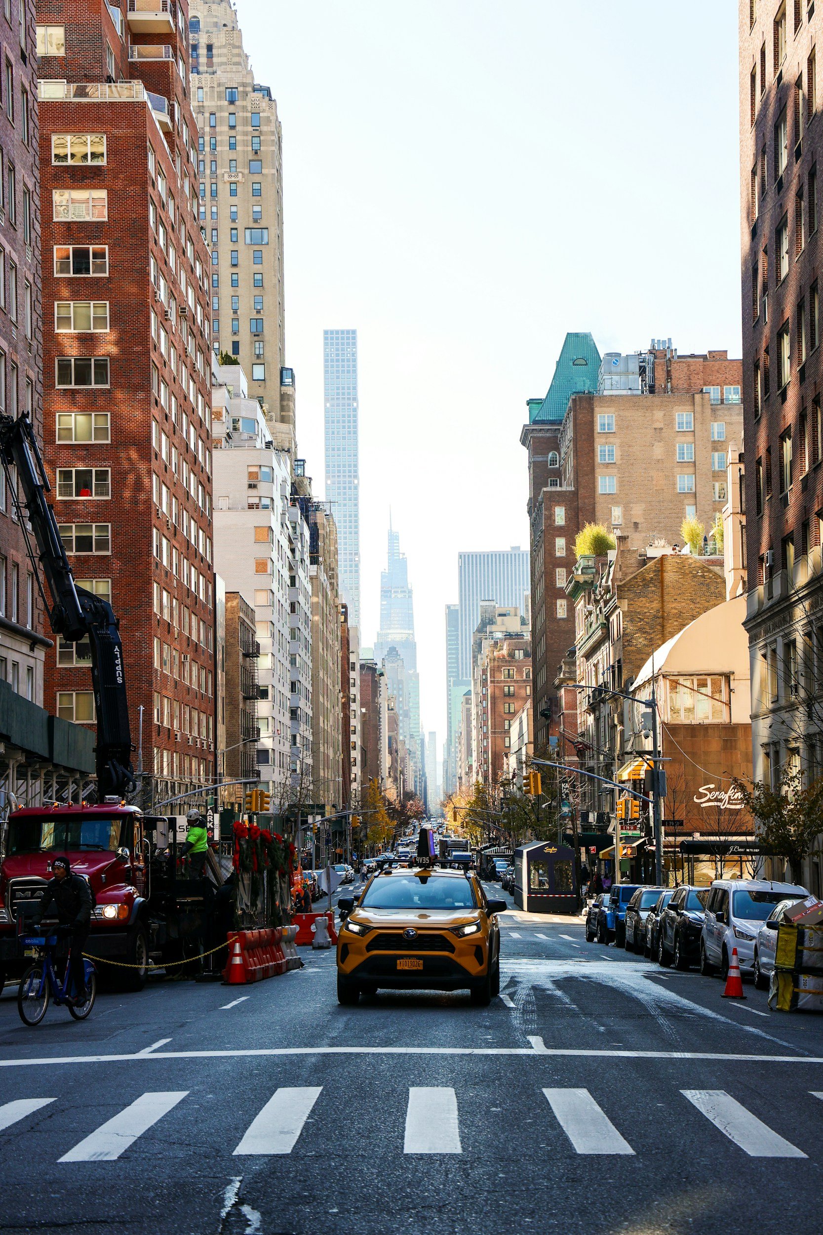 A city street lined with tall buildings and skyscrapers, with cars, pedestrians, and a cyclist on the road, and a clear sky overhead.