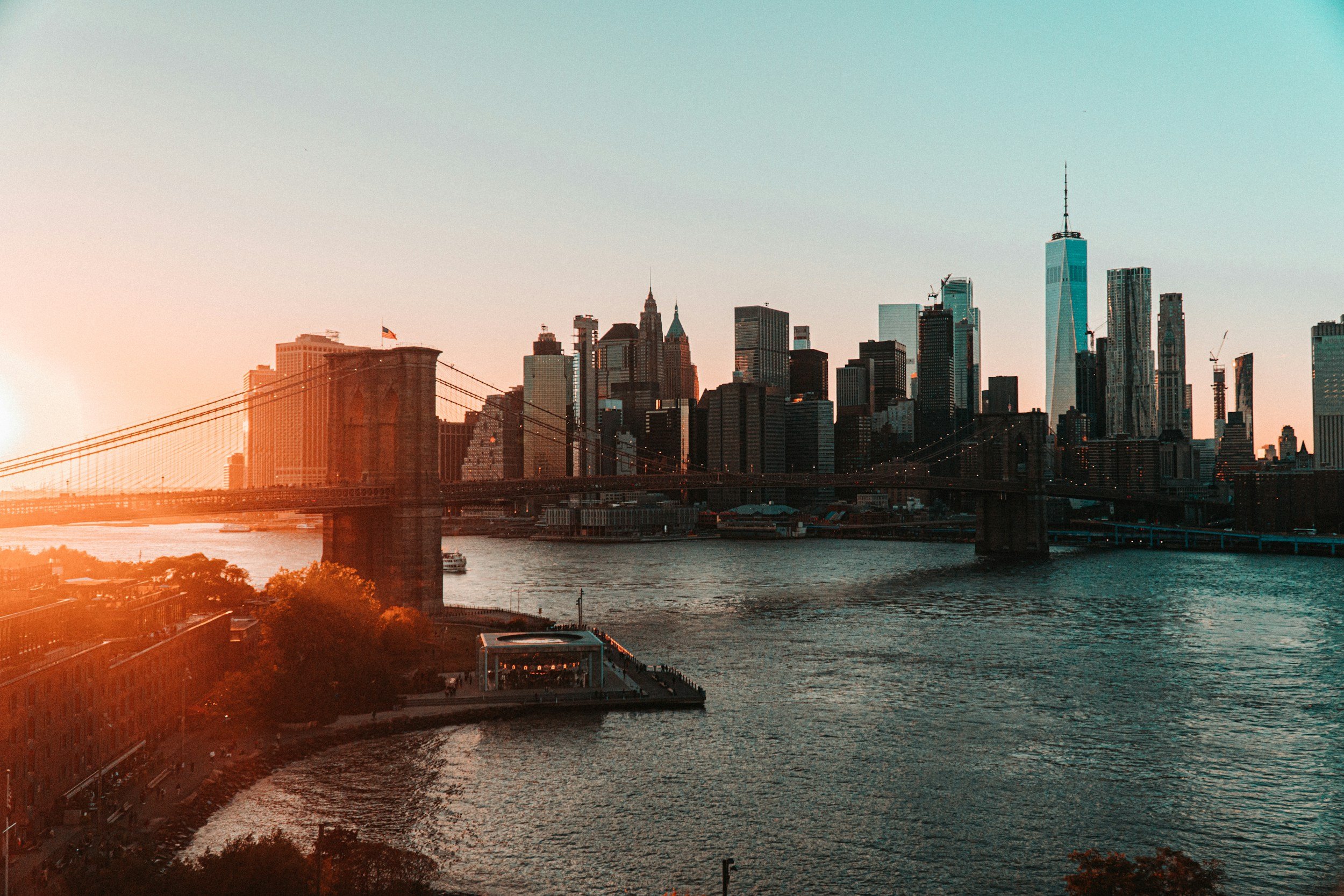 Sunset over the New York City skyline with the Brooklyn Bridge in the foreground, Brooklyn waterfront, and various skyscrapers including One World Trade Center.
