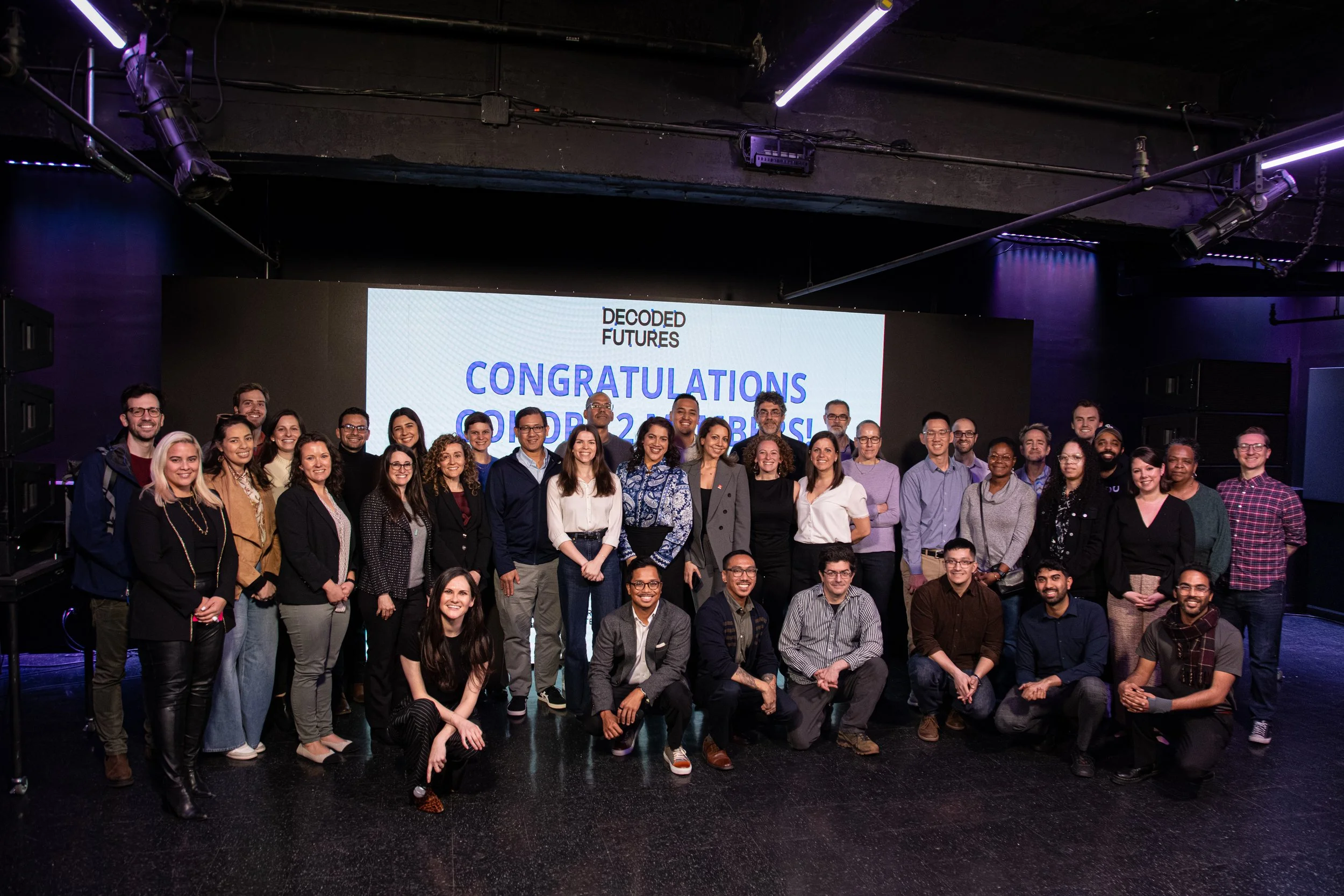 Group of diverse people gathered on stage in front of a large digital screen that says 'DECODER FUTURES' and 'CONGRATULATIONS'. They are smiling and posing for a photo at an event or ceremony.
