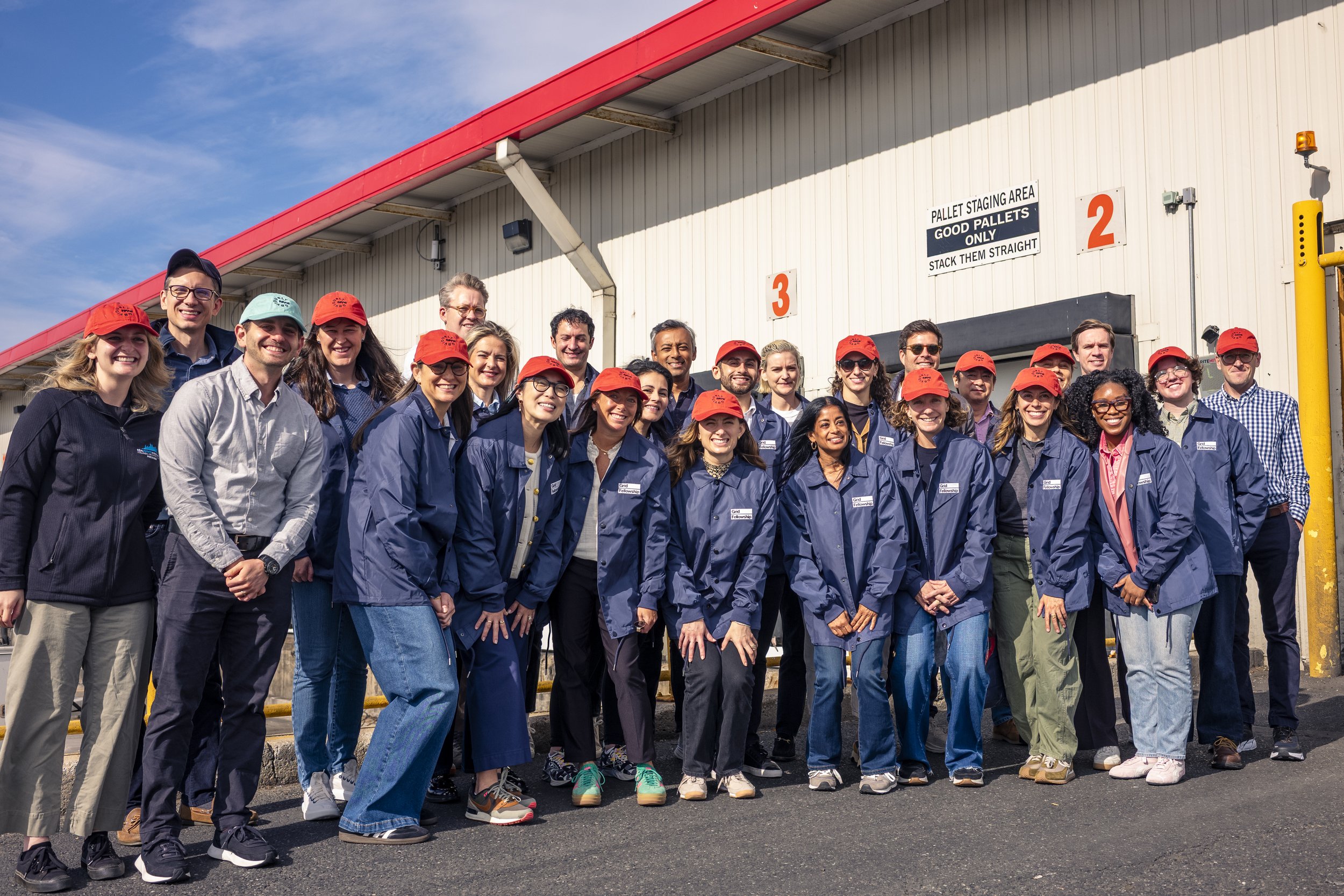 A group of diverse smiling people, some wearing red hats and blue jackets, pose outside a building under a clear sky.
