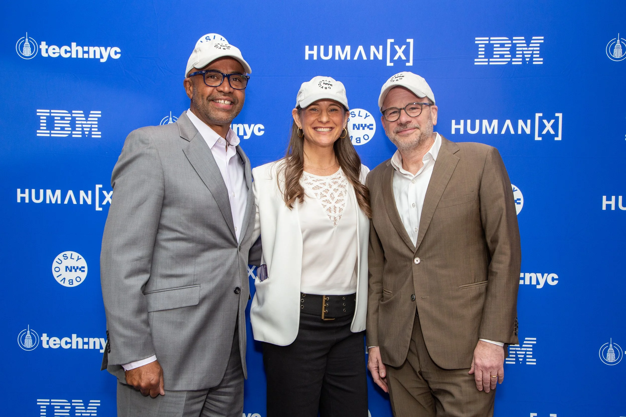 Three professionals in business attire standing on a stage with a blue backdrop displaying logos of tech companies and events, smiling for a photo.