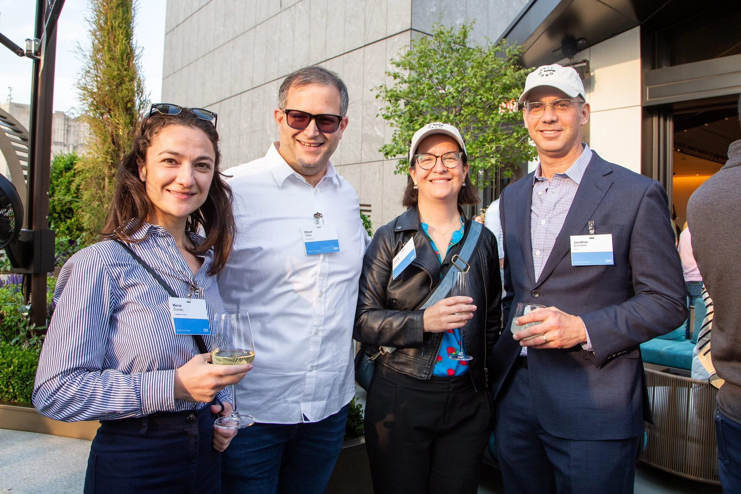 Four people smiling and holding glasses of white wine at an outdoor event, with name tags visible.