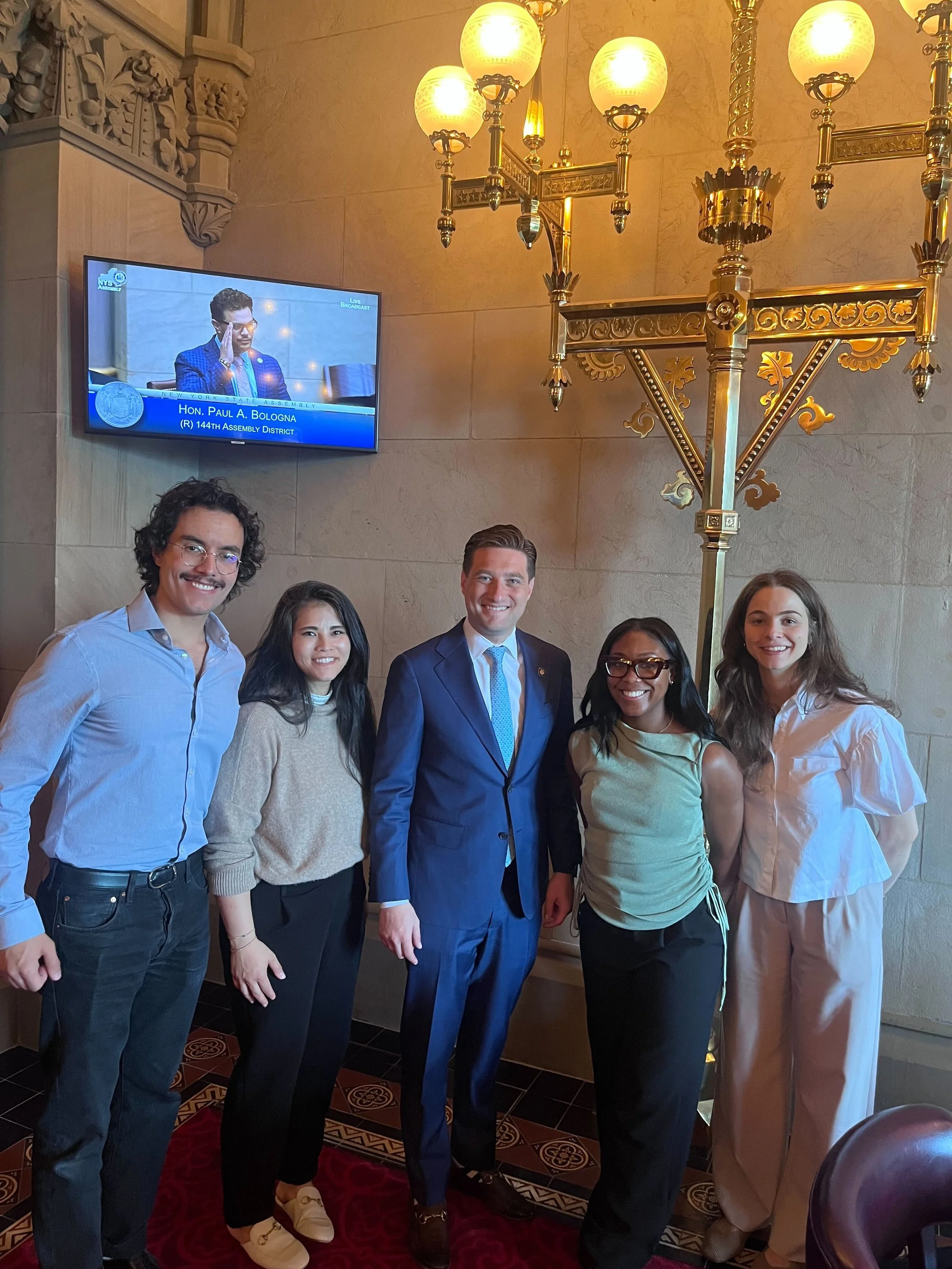 A group of five young adults standing together inside a room with ornate decor and a large chandelier, smiling at the camera.