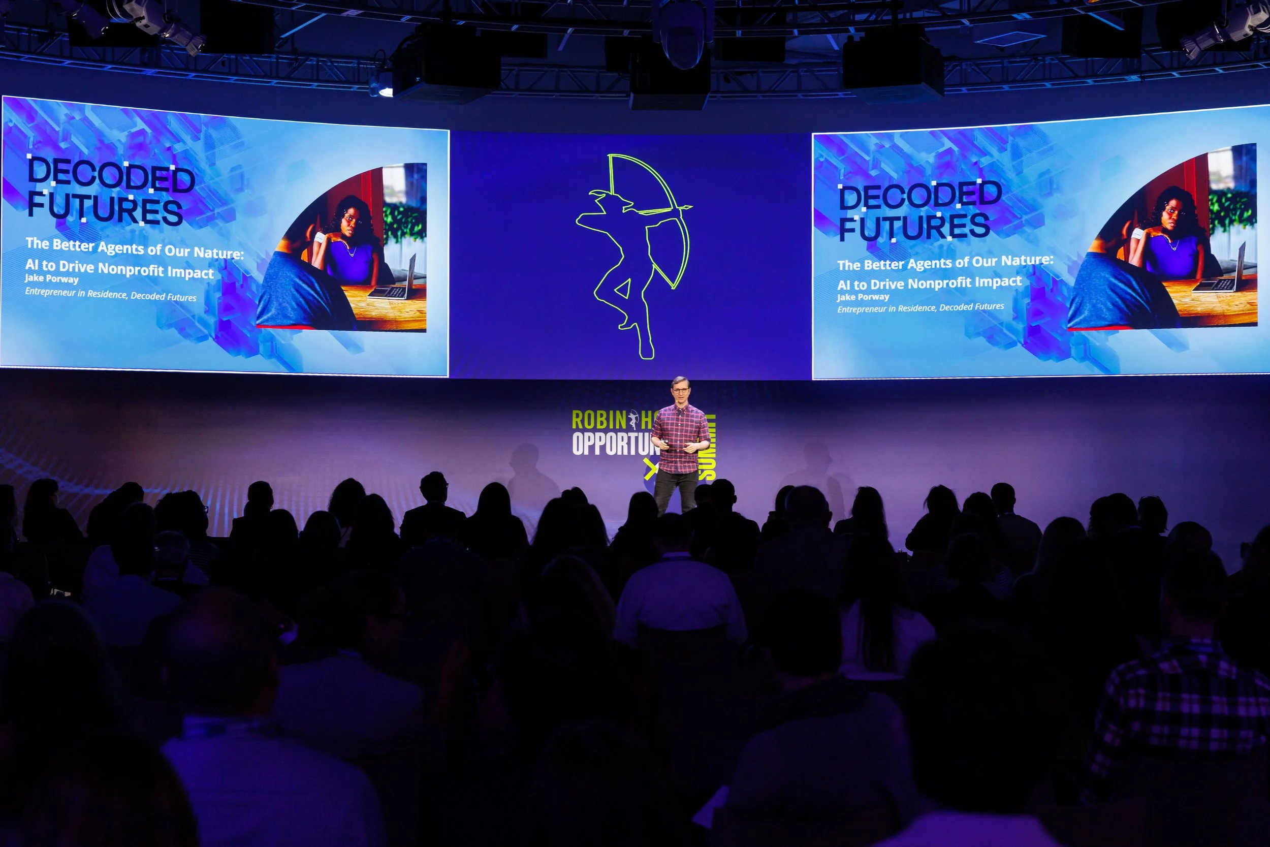 A man speaking on stage at a conference with a large screen behind him displaying a presentation titled "Decoded Futures" in blue text. The presentation slide shows an image of a woman working on a laptop and includes the text "The Better Agents of O