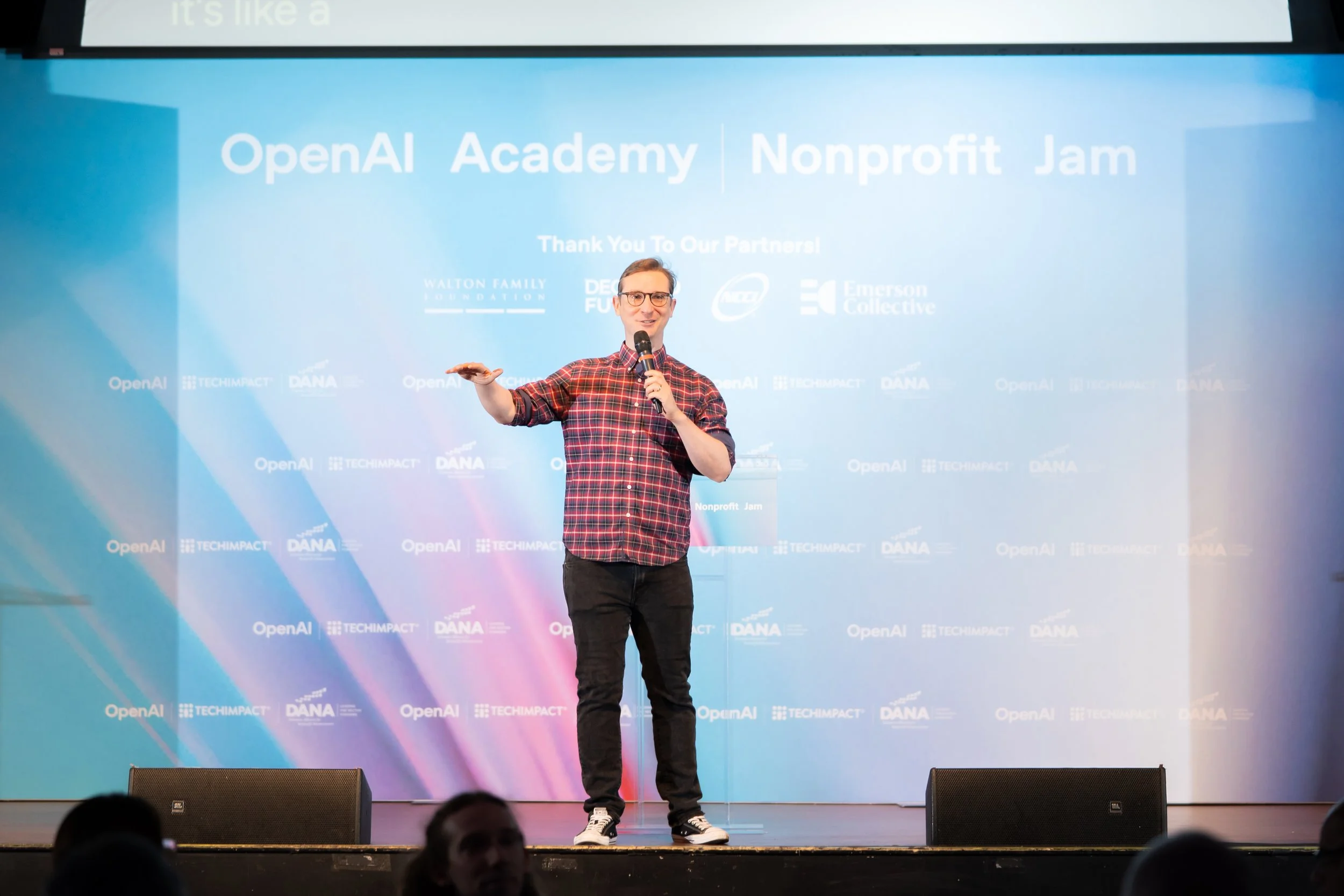 A man in a red plaid shirt and black pants is standing on a stage holding a microphone and a remote, giving a presentation at the OpenAI Academy Nonprofit Jam event, with a large blue screen behind him displaying event logos and text.