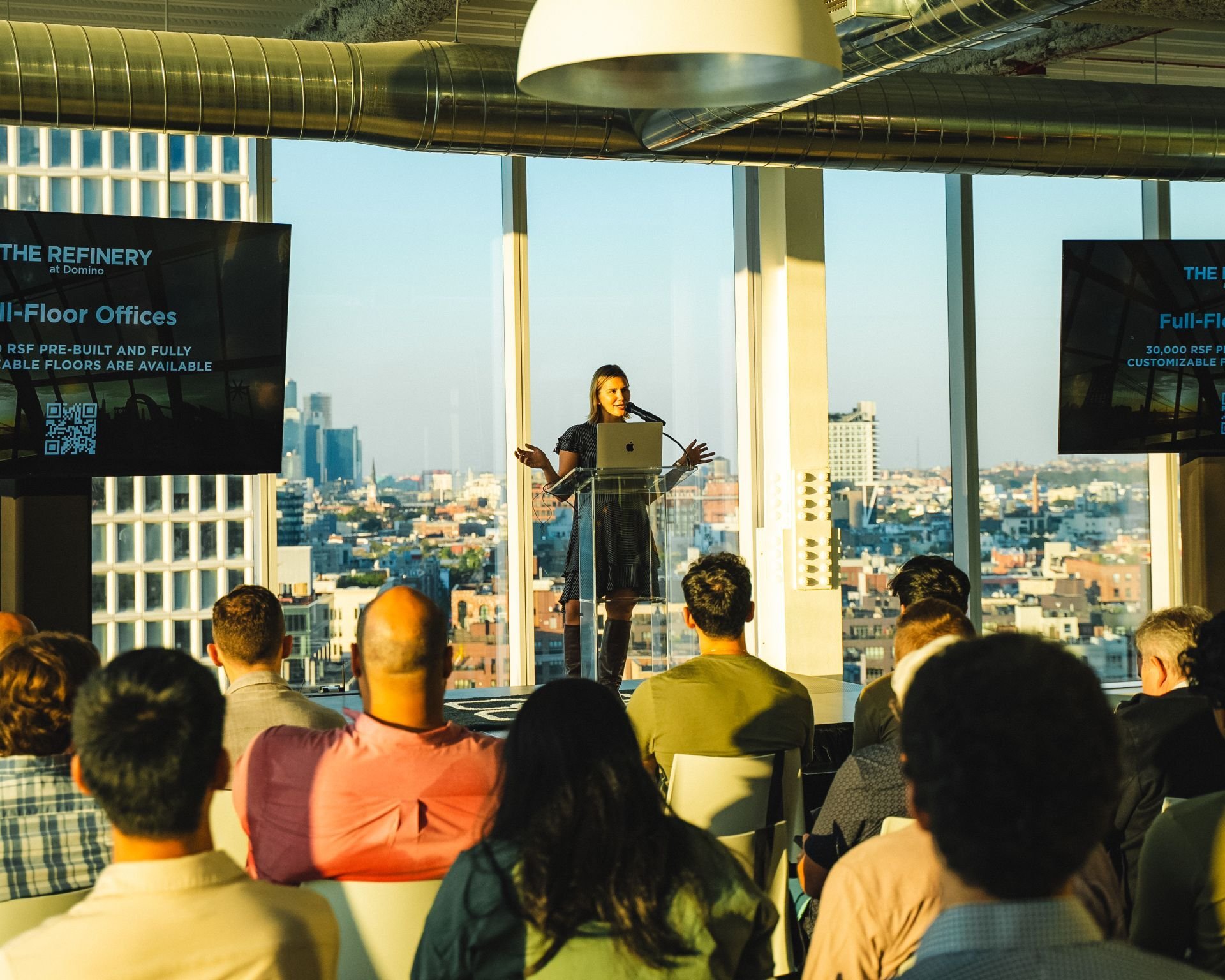 A woman giving a presentation at a conference in a high-rise building with floor-to-ceiling windows, showing a city skyline in the background. She stands behind a clear lectern with a laptop, addressing an audience seated in front of her.