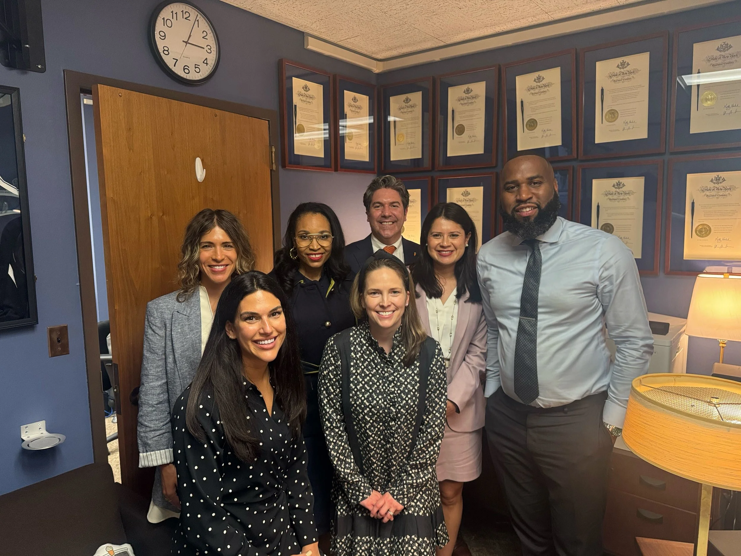 Group of seven professionals, five women and two men, gathered in an office with framed certificates on the wall, a clock, and a lamp visible.