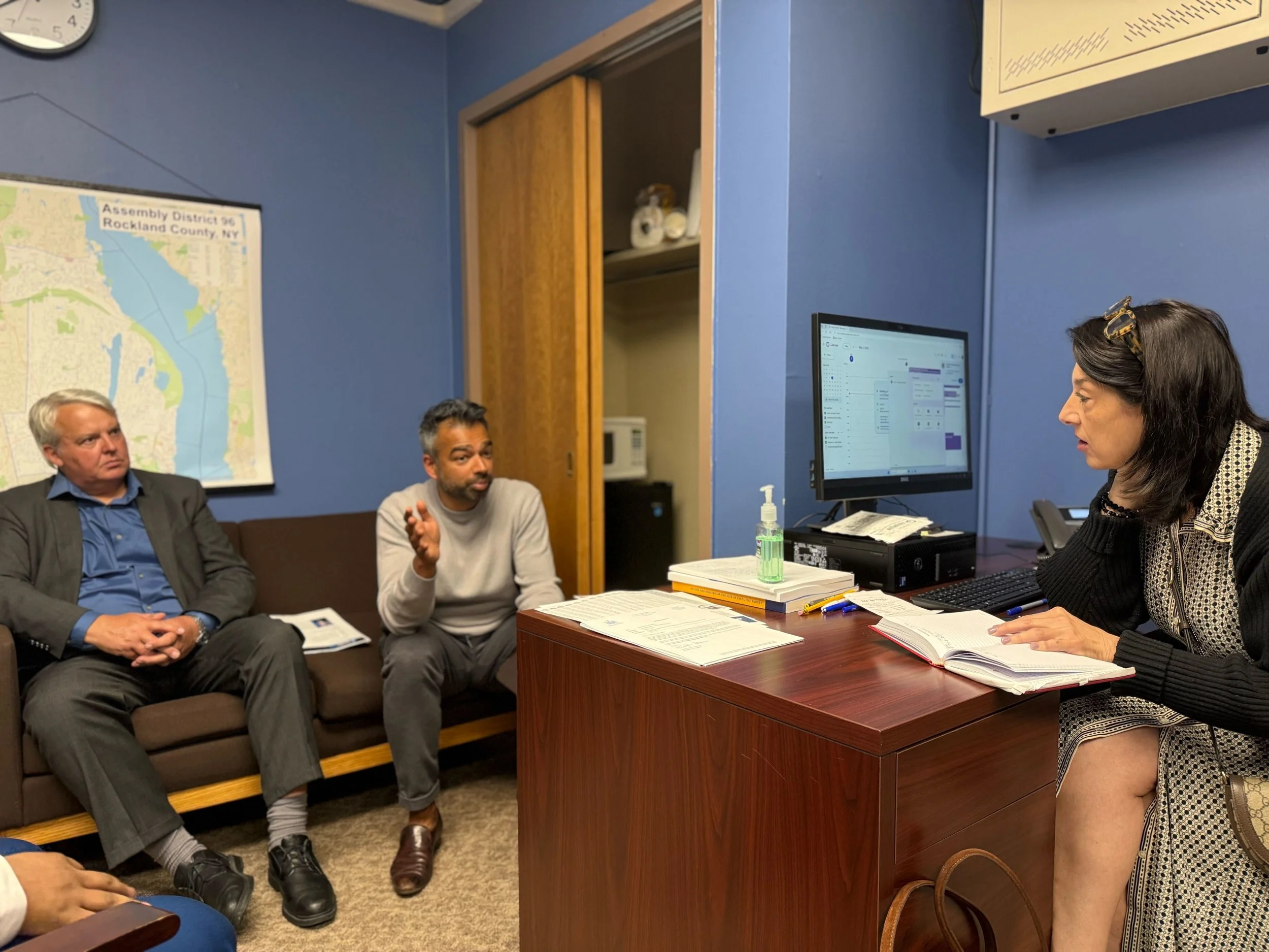 A woman with dark hair and a black sweater is sitting at a desk, speaking to two men sitting on a couch in an office. The desk has a computer, notebooks, a hand sanitizer bottle, and writing utensils. The office has blue walls, a map on the wall, and