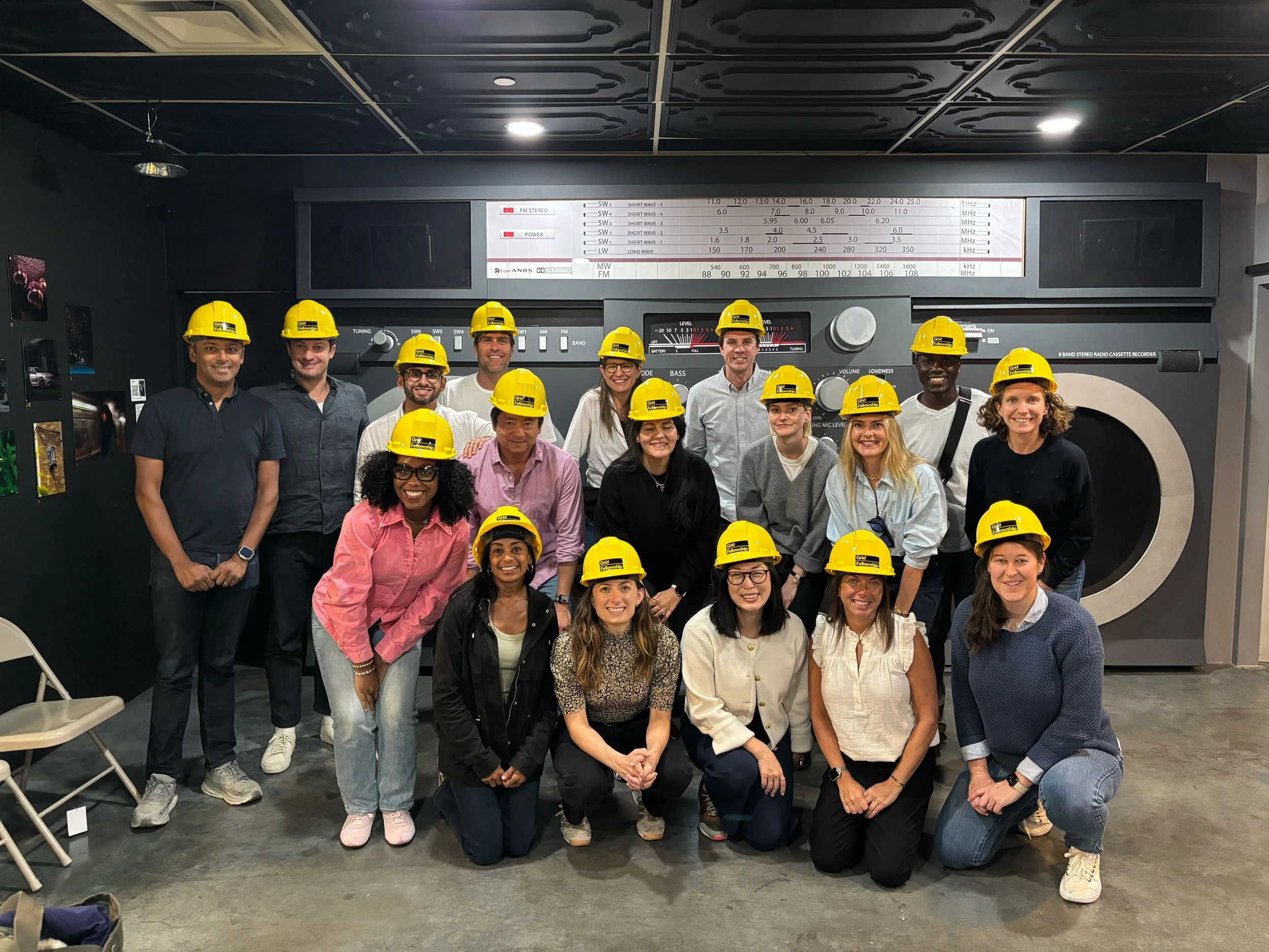 Group of people wearing yellow safety helmets standing and squatting in front of a large vintage radio or speaker in an indoor space.